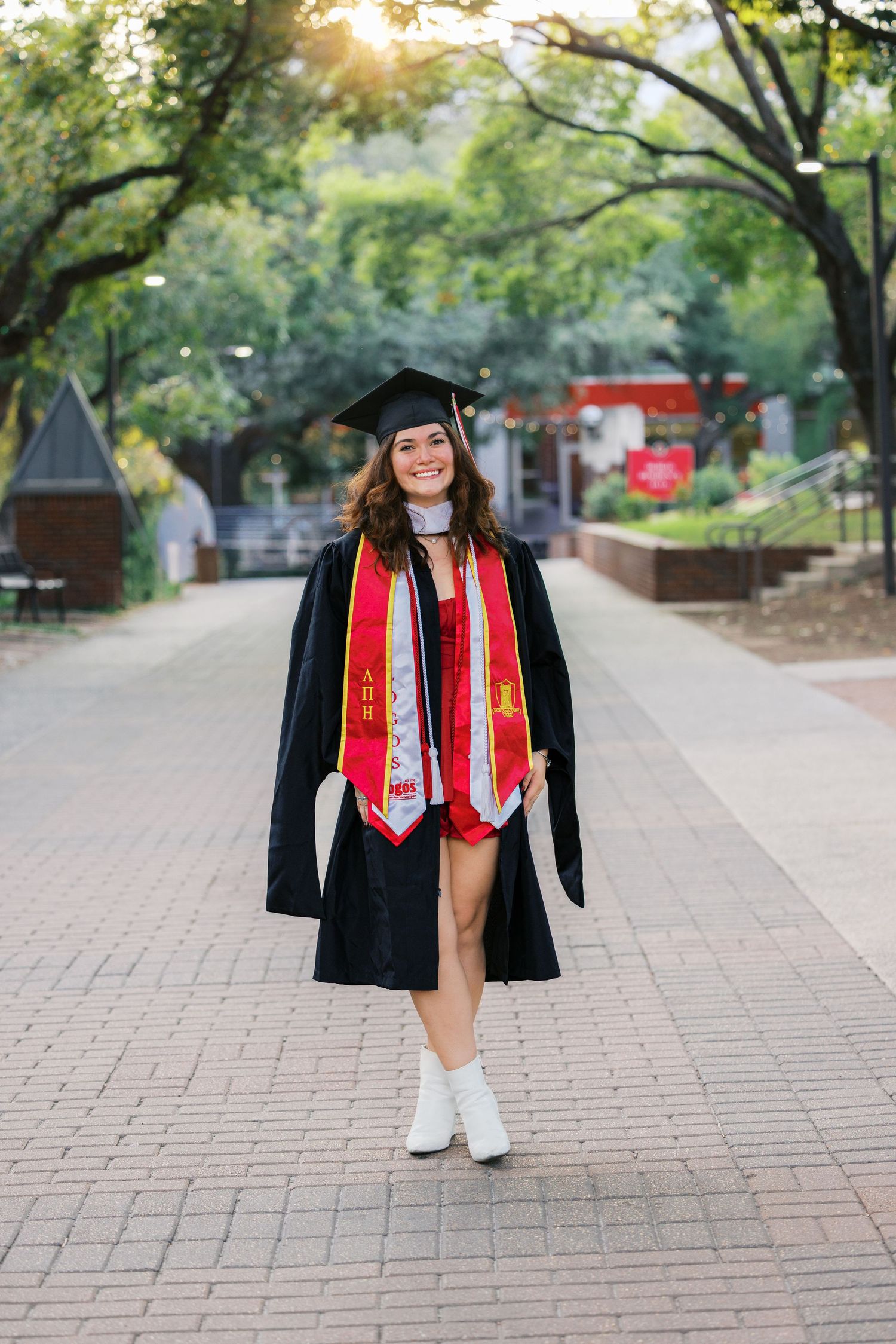 Graduate in cap and gown with red stoles standing on tree-lined campus walkway.