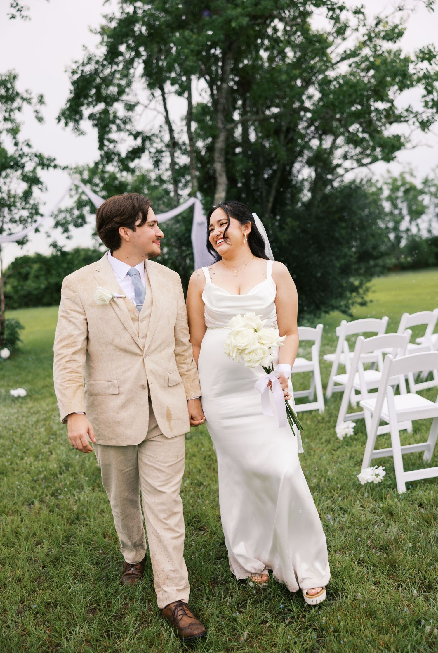 Happy bride and groom walk hand-in-hand through green outdoor wedding venue, bride holding white bouquet.