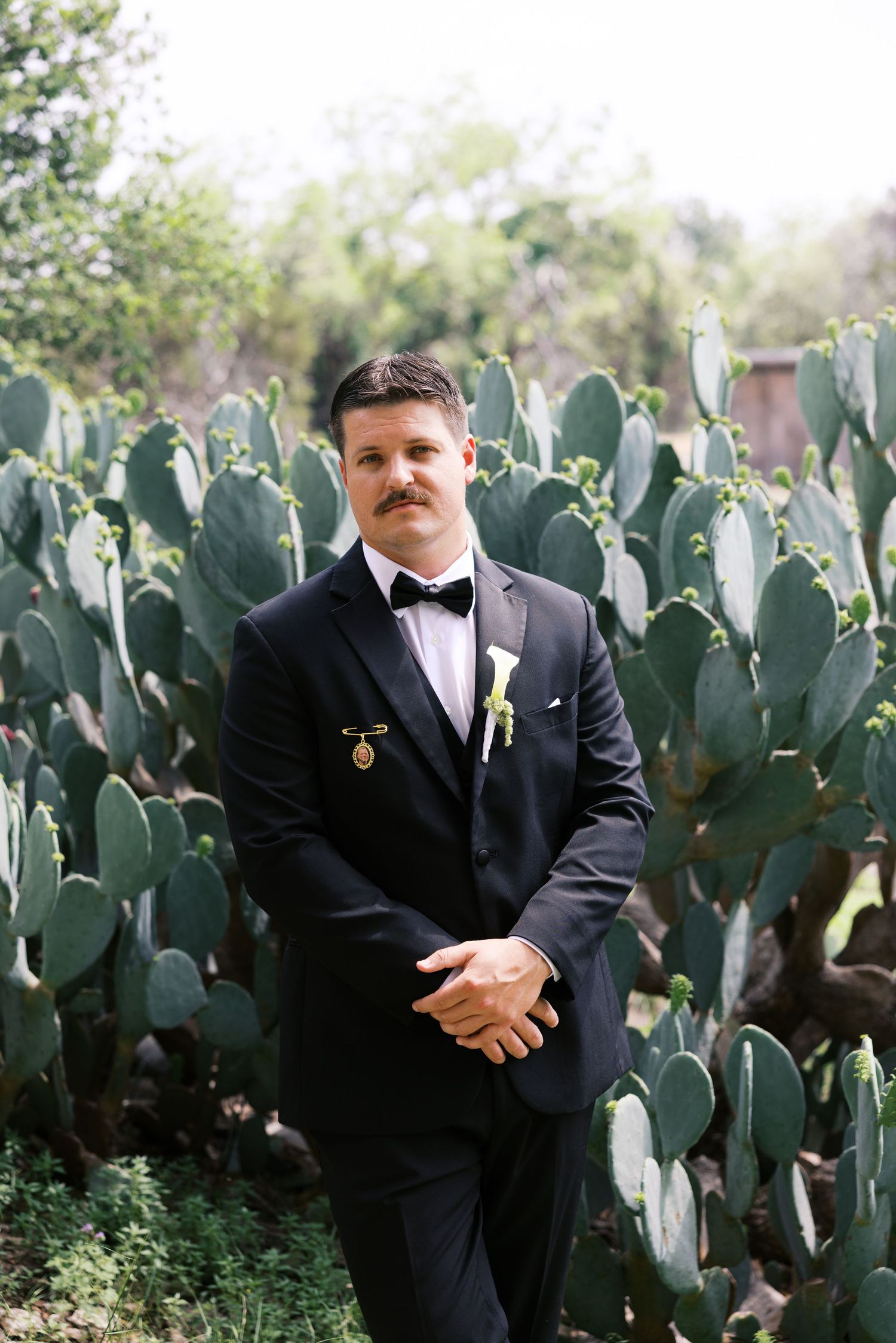 Groom in black tuxedo with bow tie and boutonniere poses in front of large green prickly pear cactus.