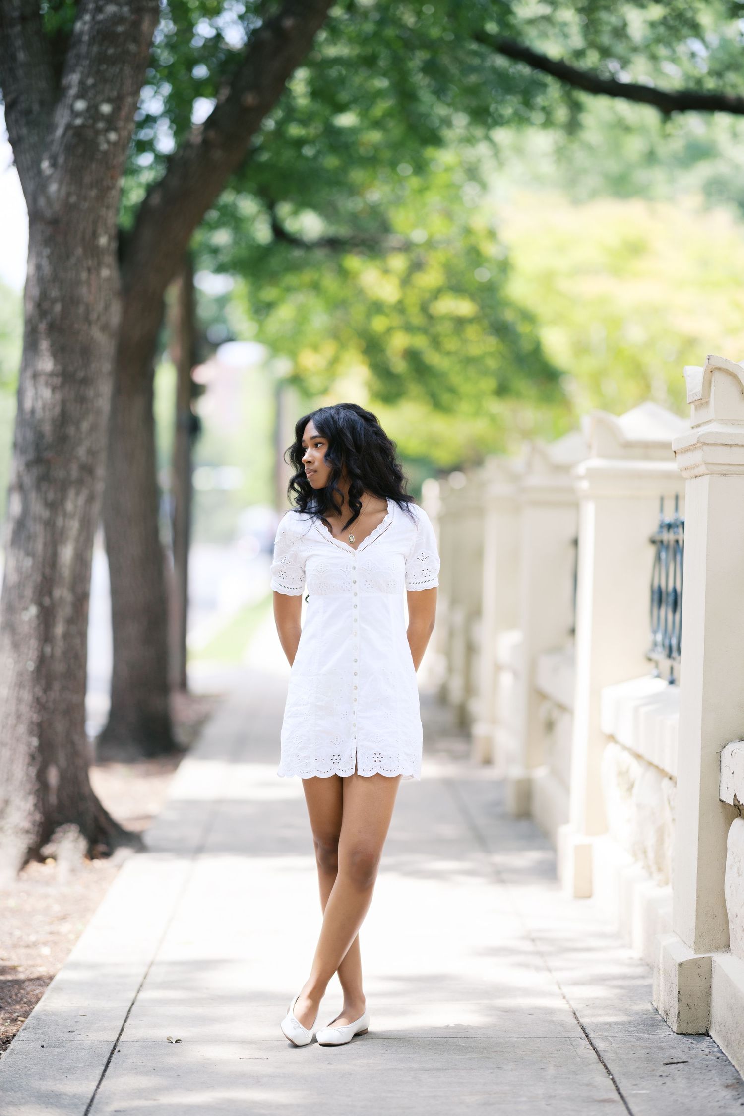 Young Black woman in white eyelet mini dress walking on sunny tree-lined sidewalk wearing white flats.
