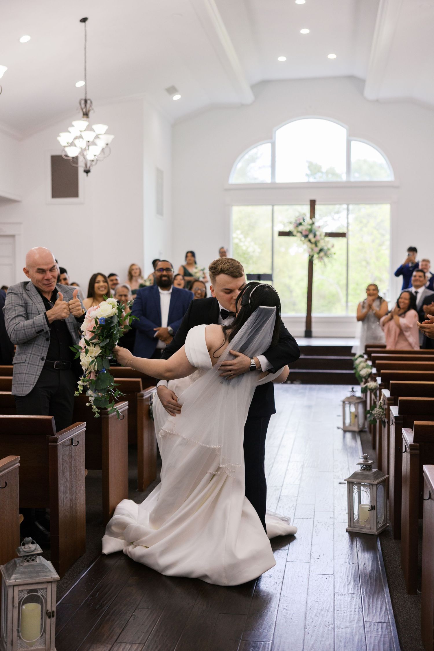 Bride and groom share a romantic dip kiss in a white chapel aisle surrounded by guests during their wedding ceremony.
