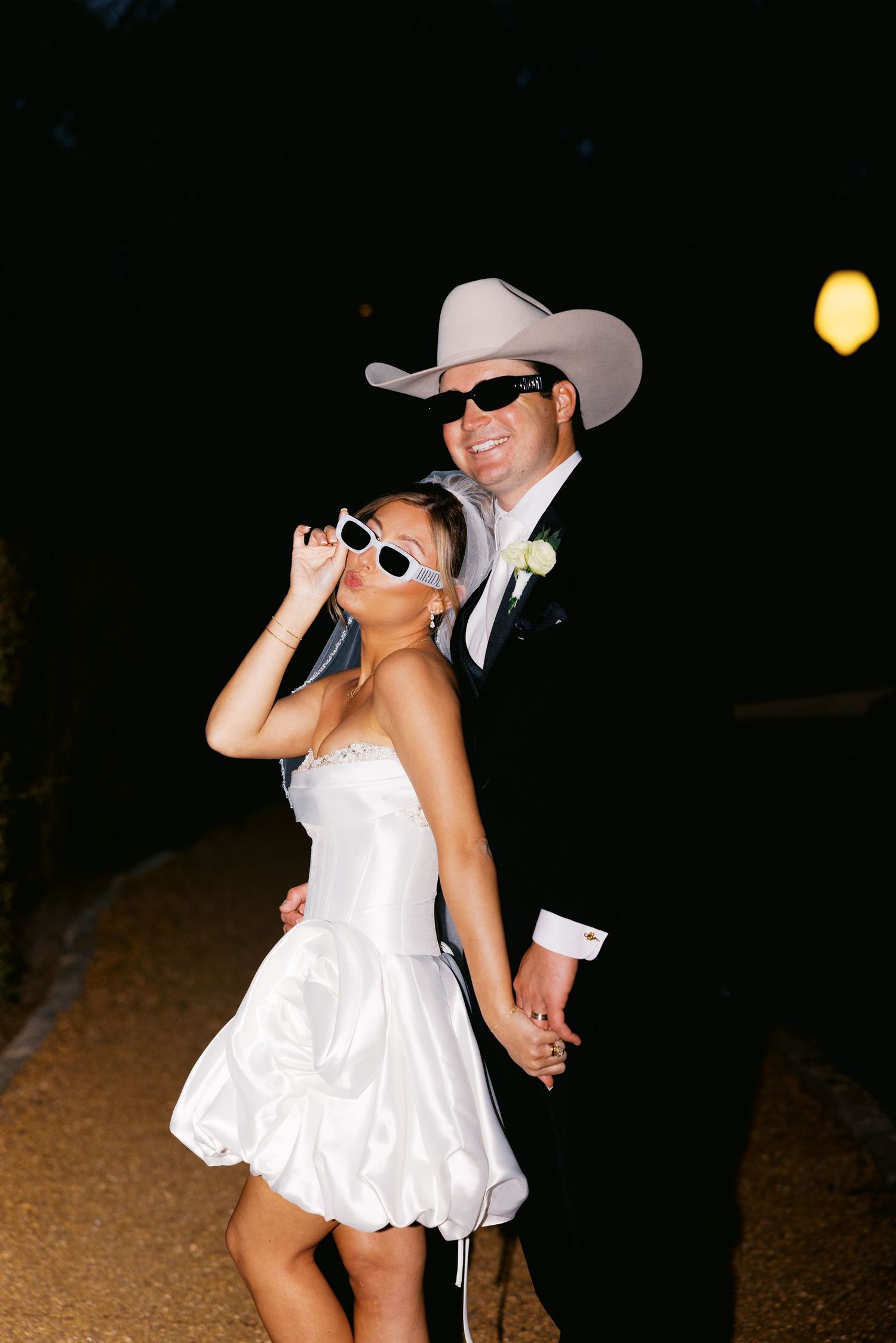 Bride in white mini dress and groom in black tuxedo and cowboy hat pose with sunglasses at night.