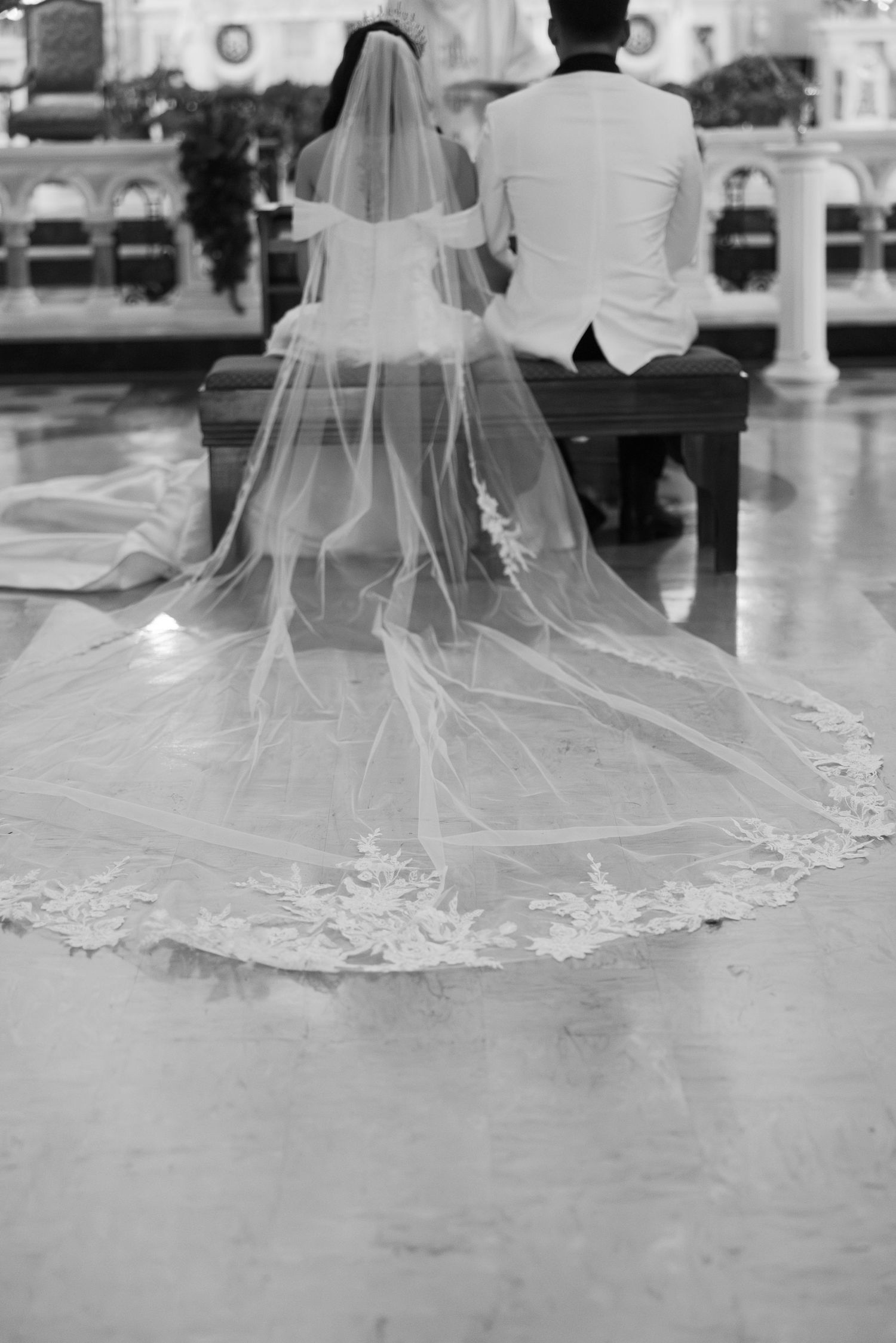 A long lace wedding veil trailing behind a seated couple during a church ceremony.