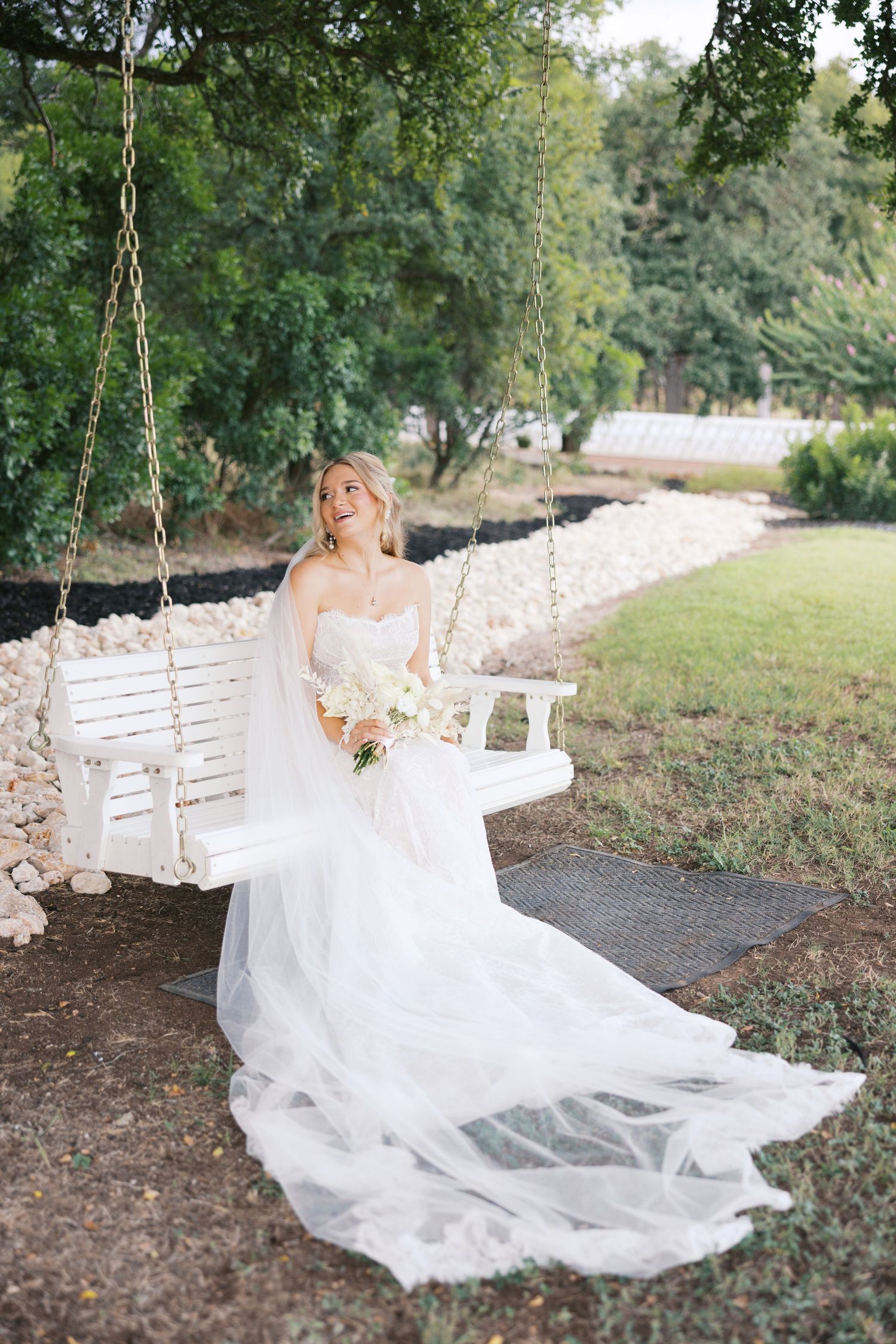 Bride in flowing white gown poses on rustic garden swing with natural backdrop.