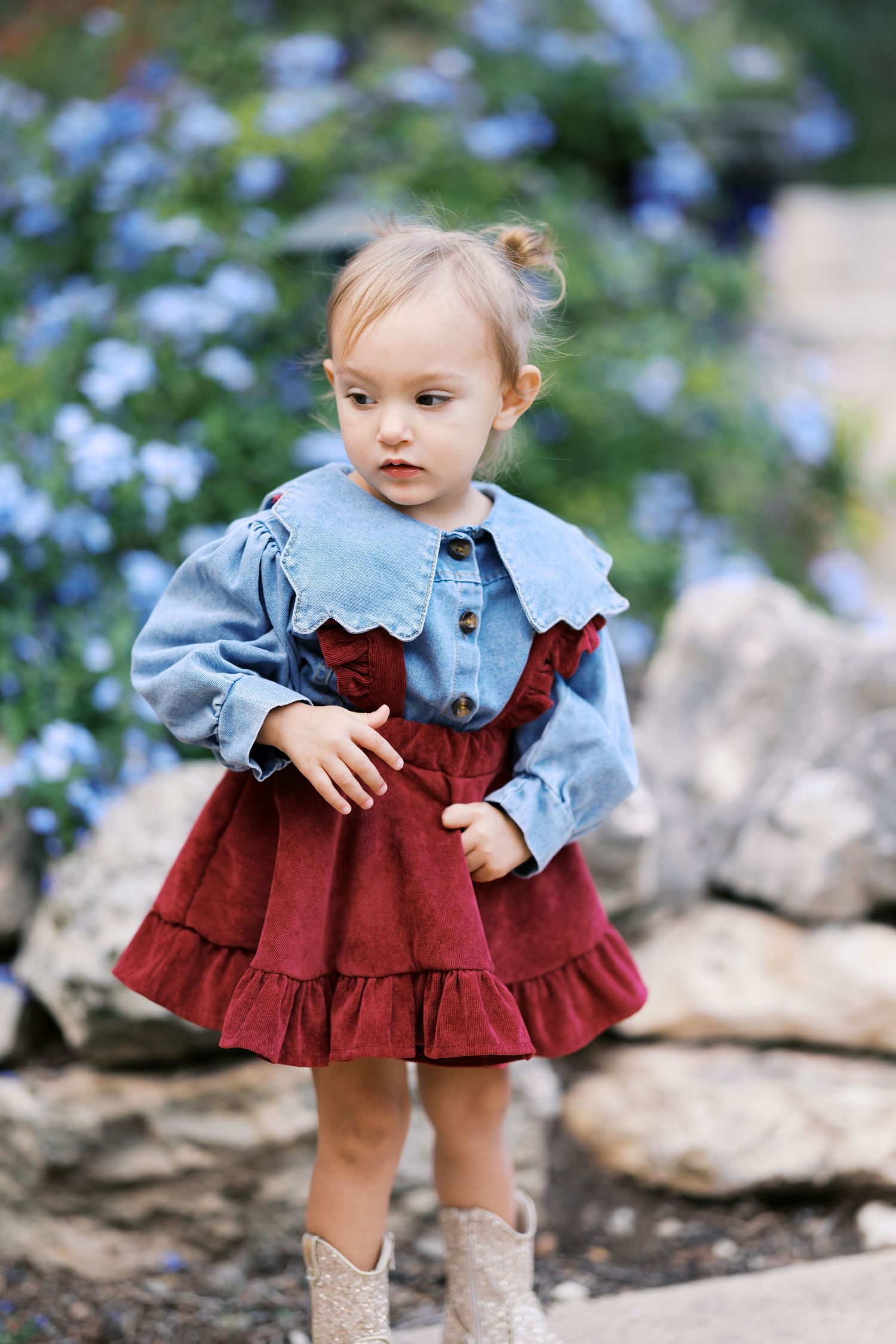 A small child wearing a burgundy dress with blue ruffled collar stands on rocks surrounded by blue flowers.
