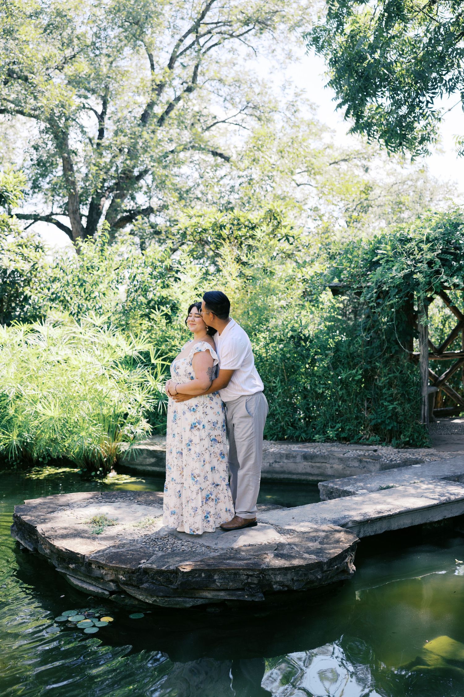 A romantic moment captured on a stone platform overlooking a serene pond surrounded by lush trees.