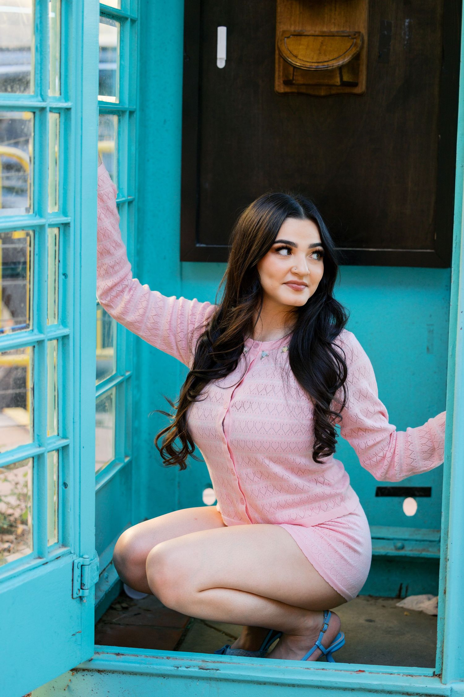 A casual pose inside a turquoise phone booth wearing pink attire.