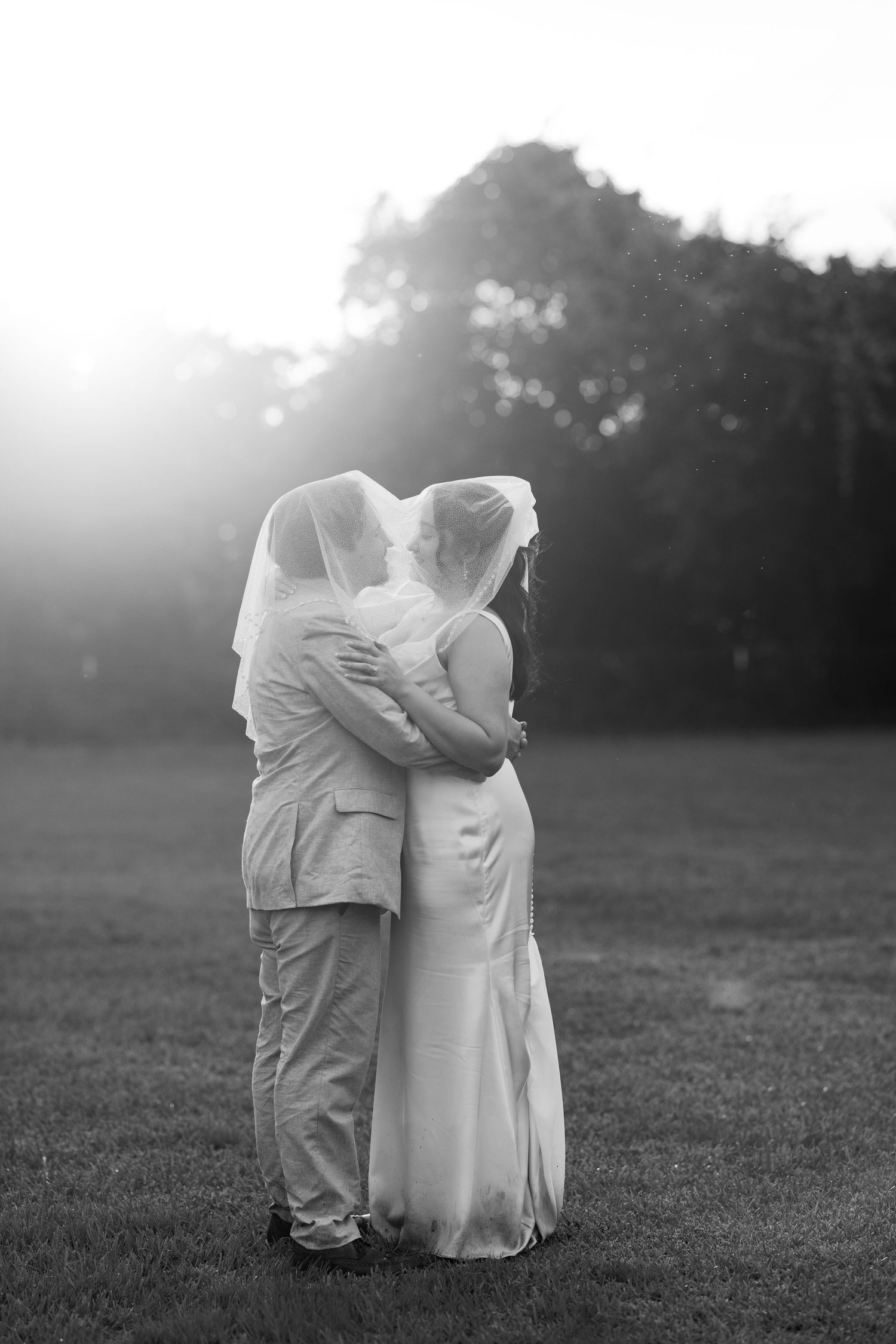 Black and white romantic photo of newlyweds embracing under veil at sunset in an open field.