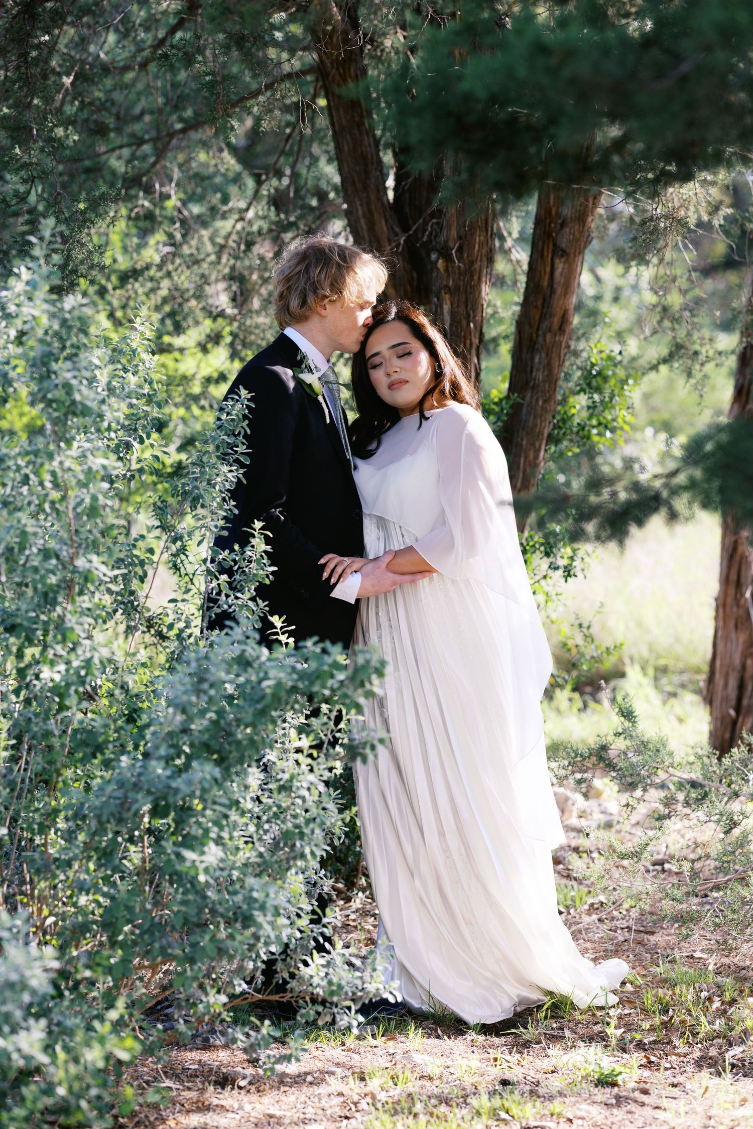 Couple in wedding attire embracing in a lush forest, bride in white gown and groom in black suit.