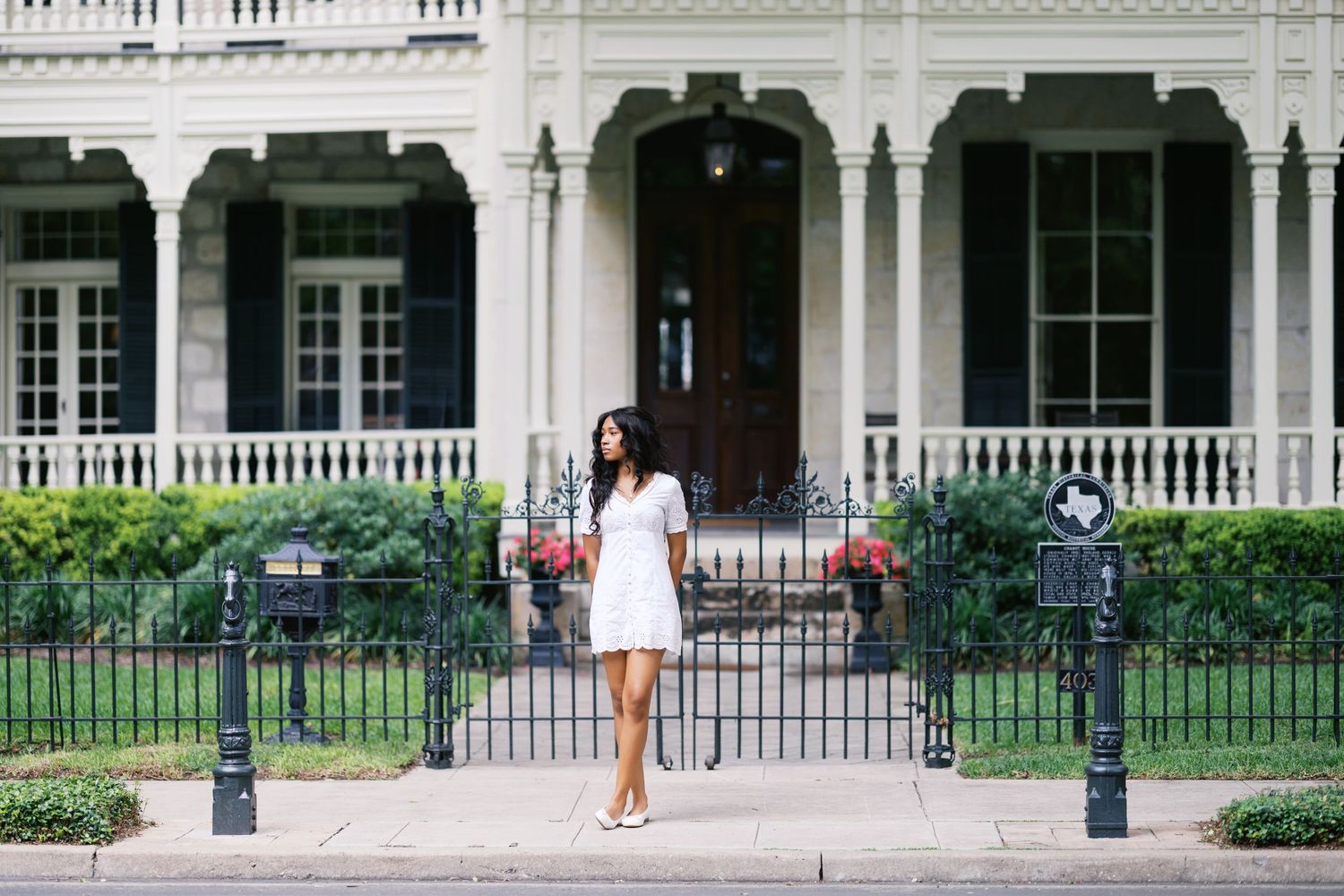 Woman in white dress standing in front of elegant Victorian home with ornate iron fence in New Orleans.