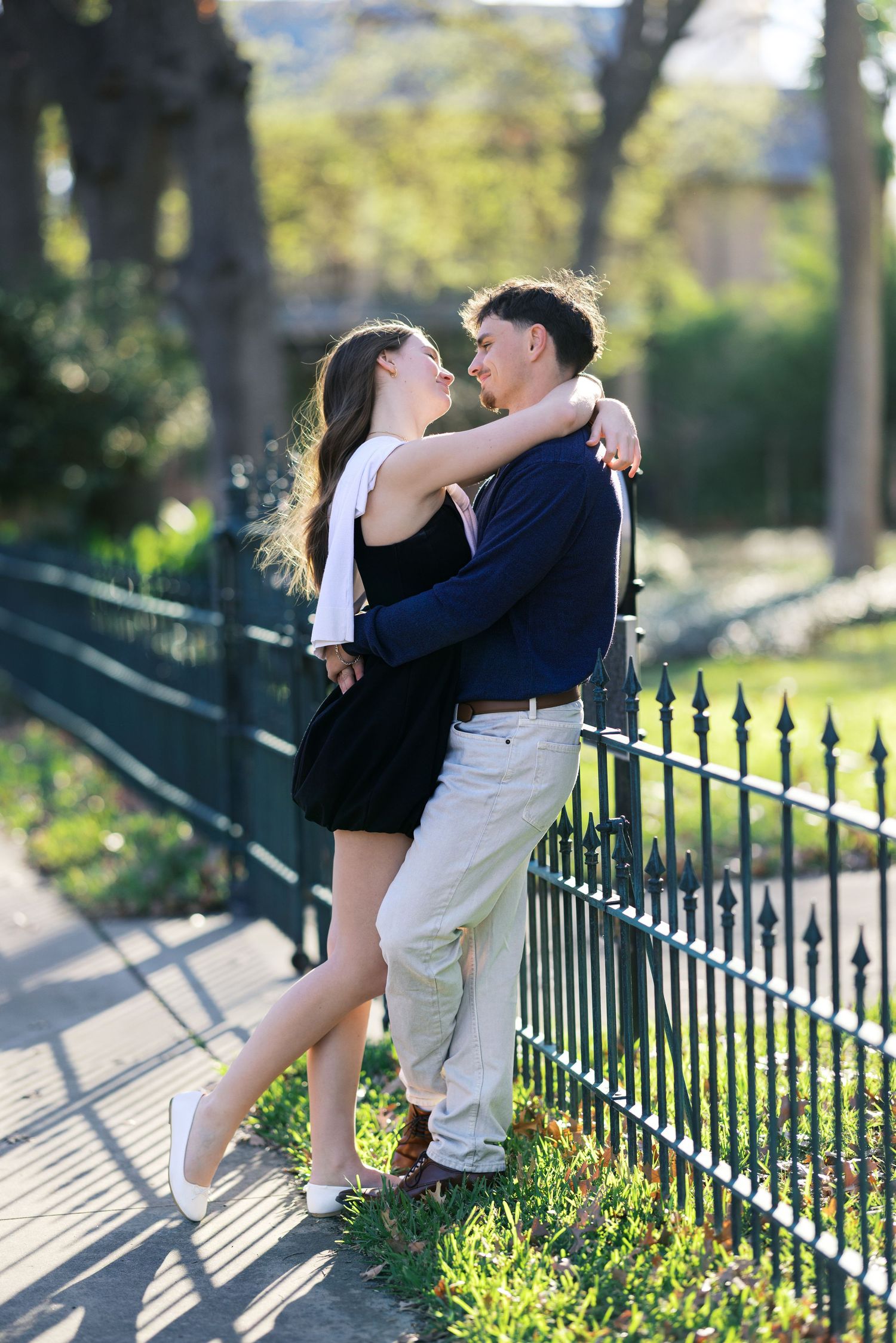 Young couple embracing and gazing at each other by ornate iron fence in sunny park setting.