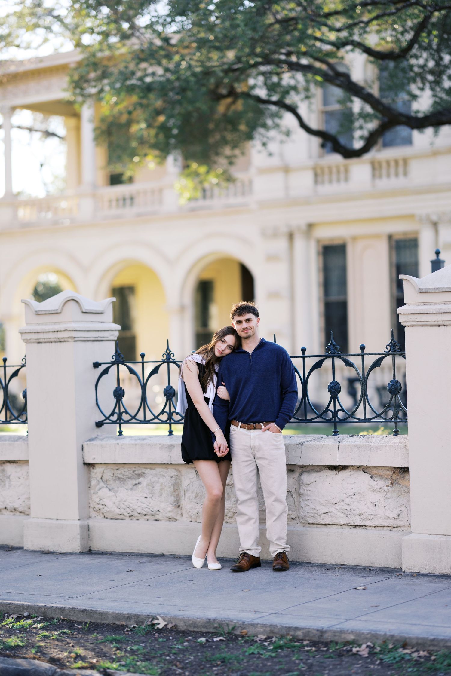 Young couple posing together by an ornate iron fence in front of a classic historic building.