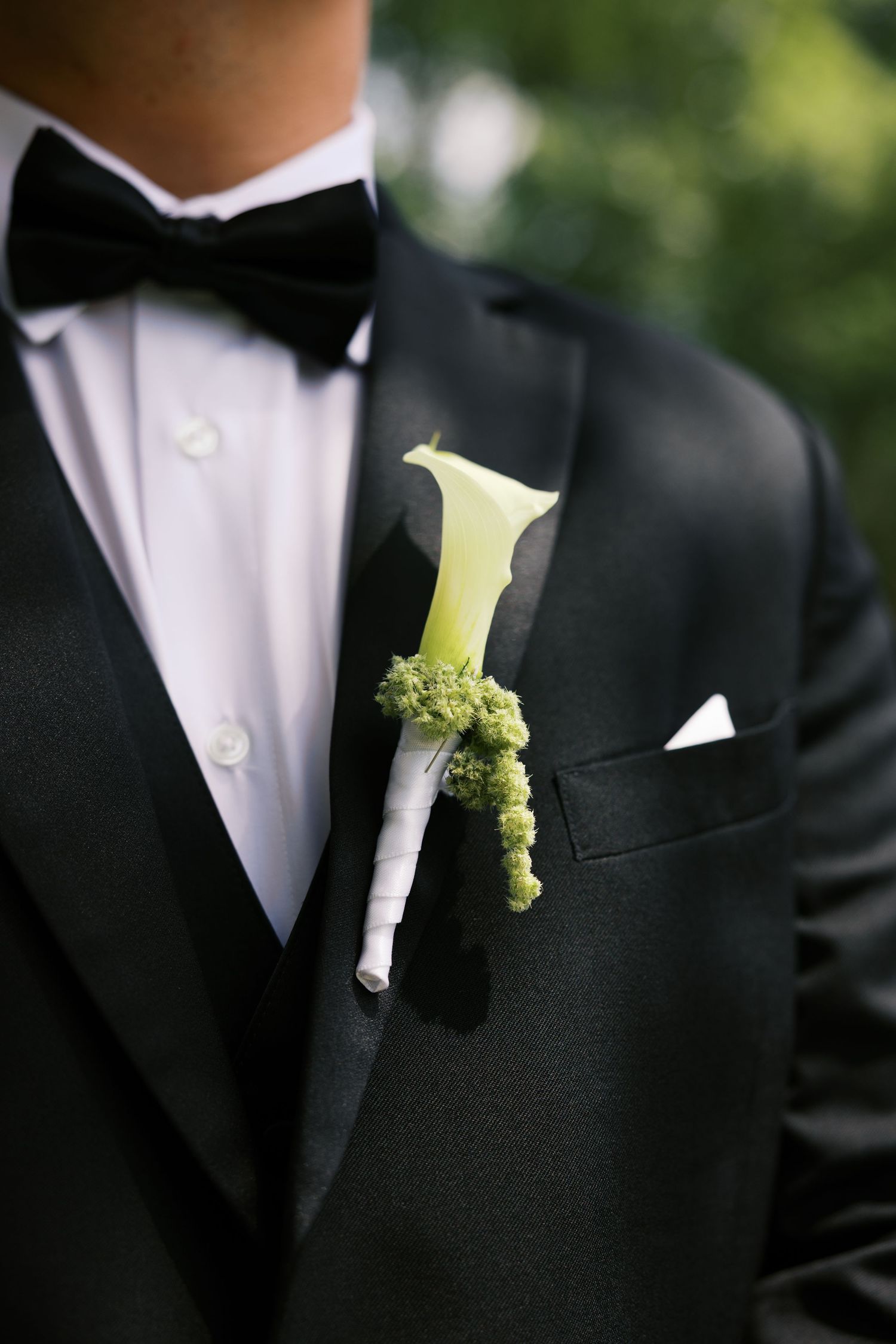 Groom in black tuxedo with bow tie wearing a white calla lily and green boutonniere on his lapel.