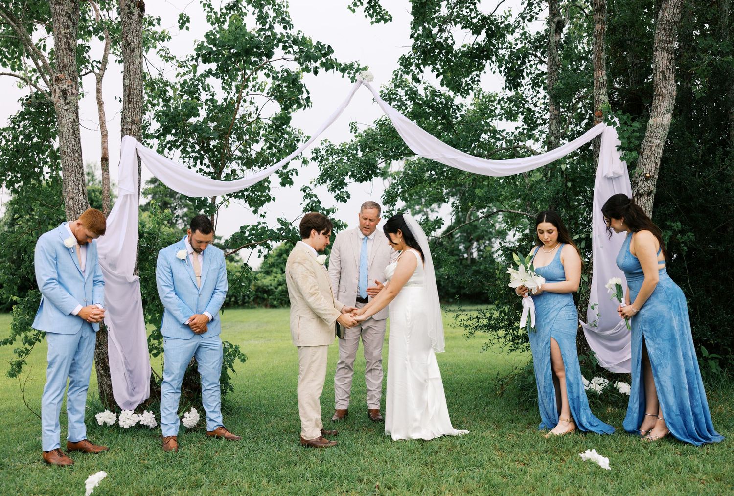 Outdoor wedding ceremony on green lawn with couple, officiant, groomsmen in blue suits, and bridesmaids in blue dresses.