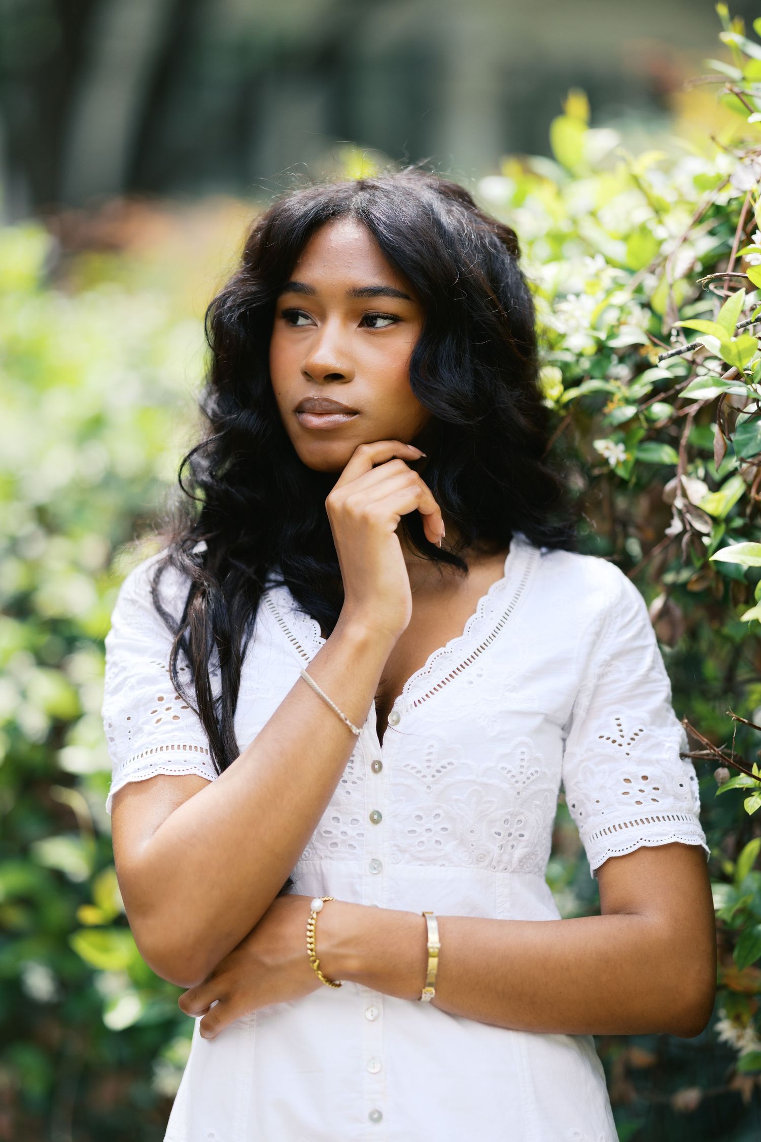 Young Black woman in white eyelet dress with gold bracelets posing thoughtfully beside lush green bushes.