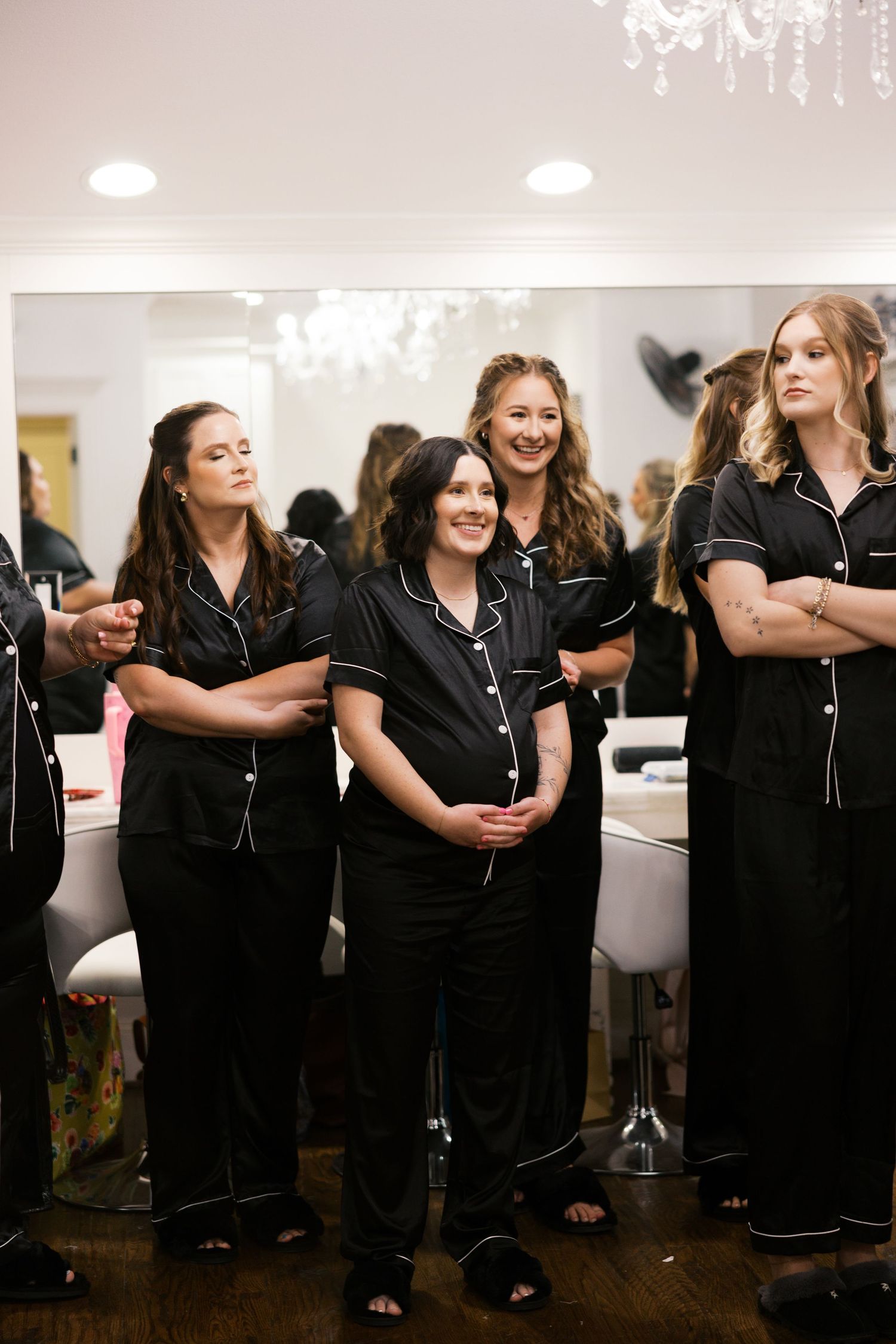 Bridesmaids in matching black satin pajamas with white piping smile together in a bridal suite before the wedding.