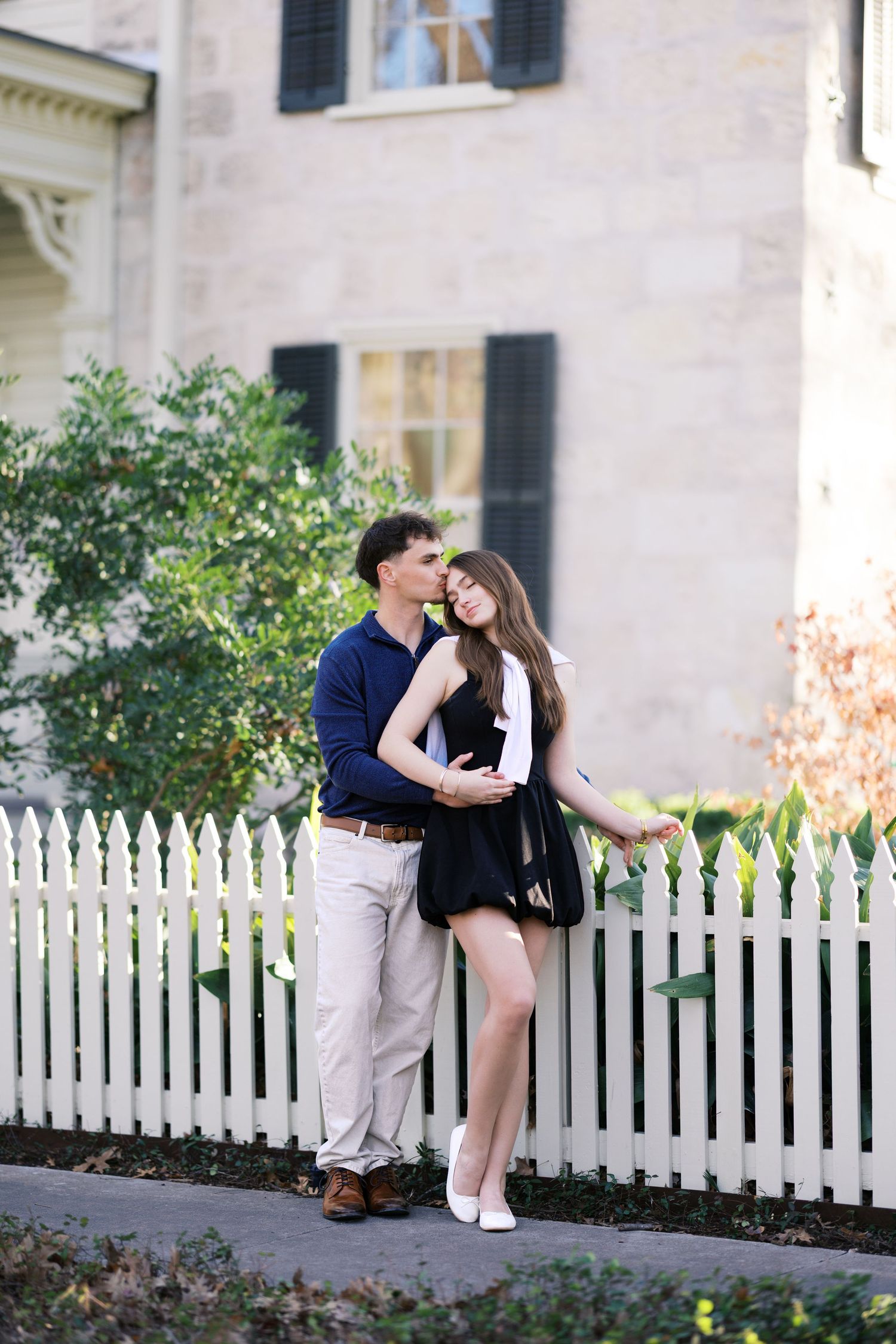 Romantic couple embracing by a white picket fence in front of a charming historic stone home.