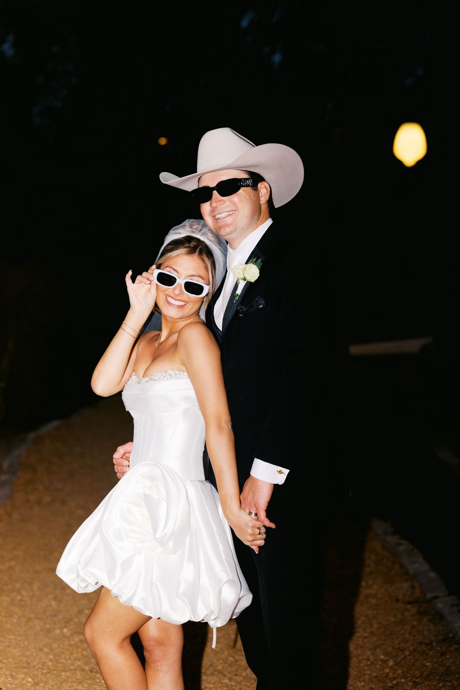 Stylish newlyweds pose at night wearing matching sunglasses, groom in cowboy hat, bride in short white dress.