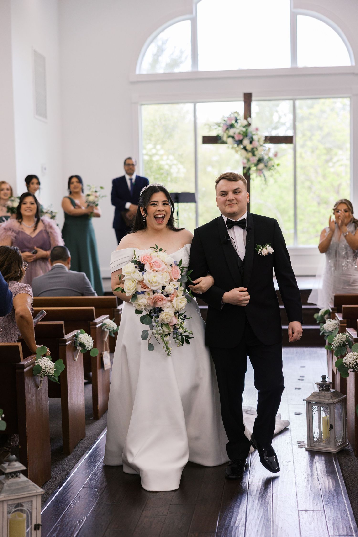 Joyful bride and groom walking down the aisle after wedding ceremony in a bright white chapel with floral decorations.