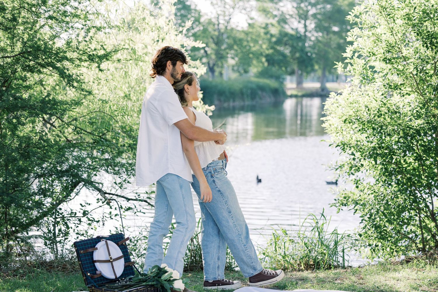 A couple standing together by a lake with a picnic setup on a white blanket.