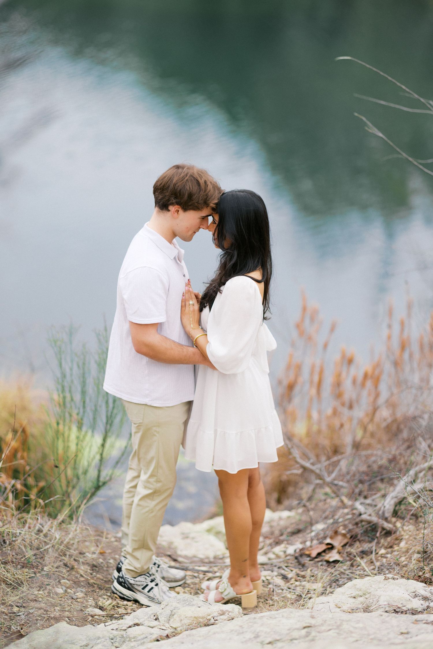 Two people in light colored clothing embrace while standing on rocky lakeside terrain.
