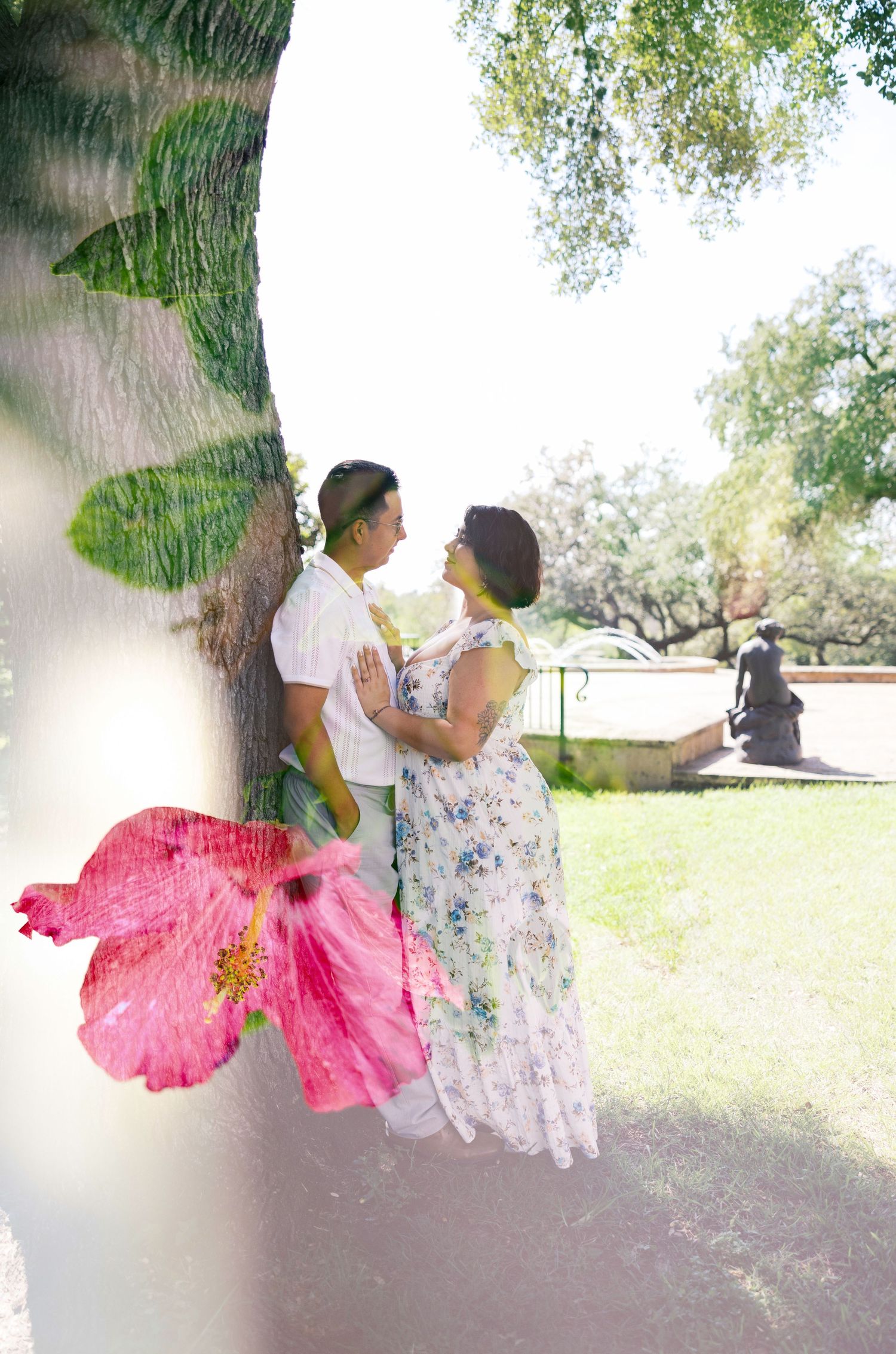 A couple embraces near a tree with a vibrant pink hibiscus flower in the foreground.