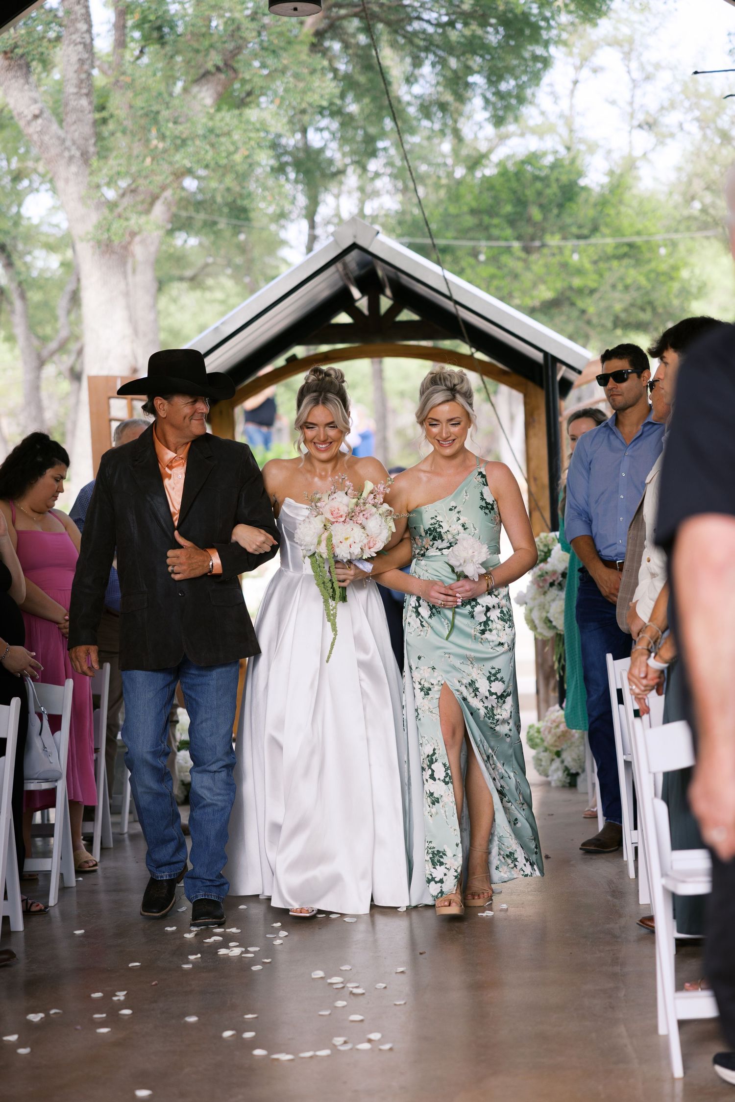 Bride walking down the aisle escorted by two people at an outdoor wedding ceremony.