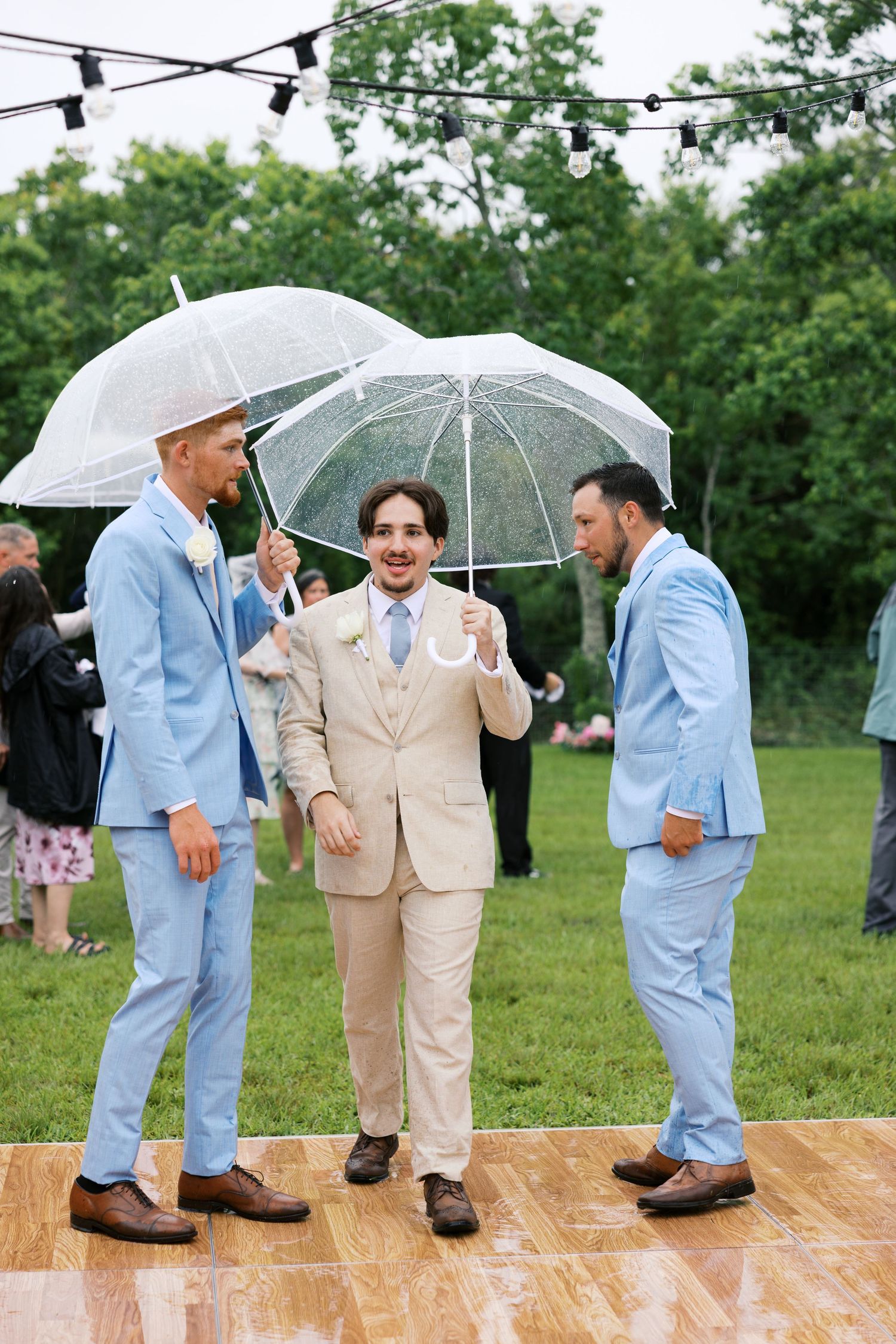 Groom in beige suit holds clear umbrella with groomsmen in blue suits during rainy outdoor wedding ceremony.
