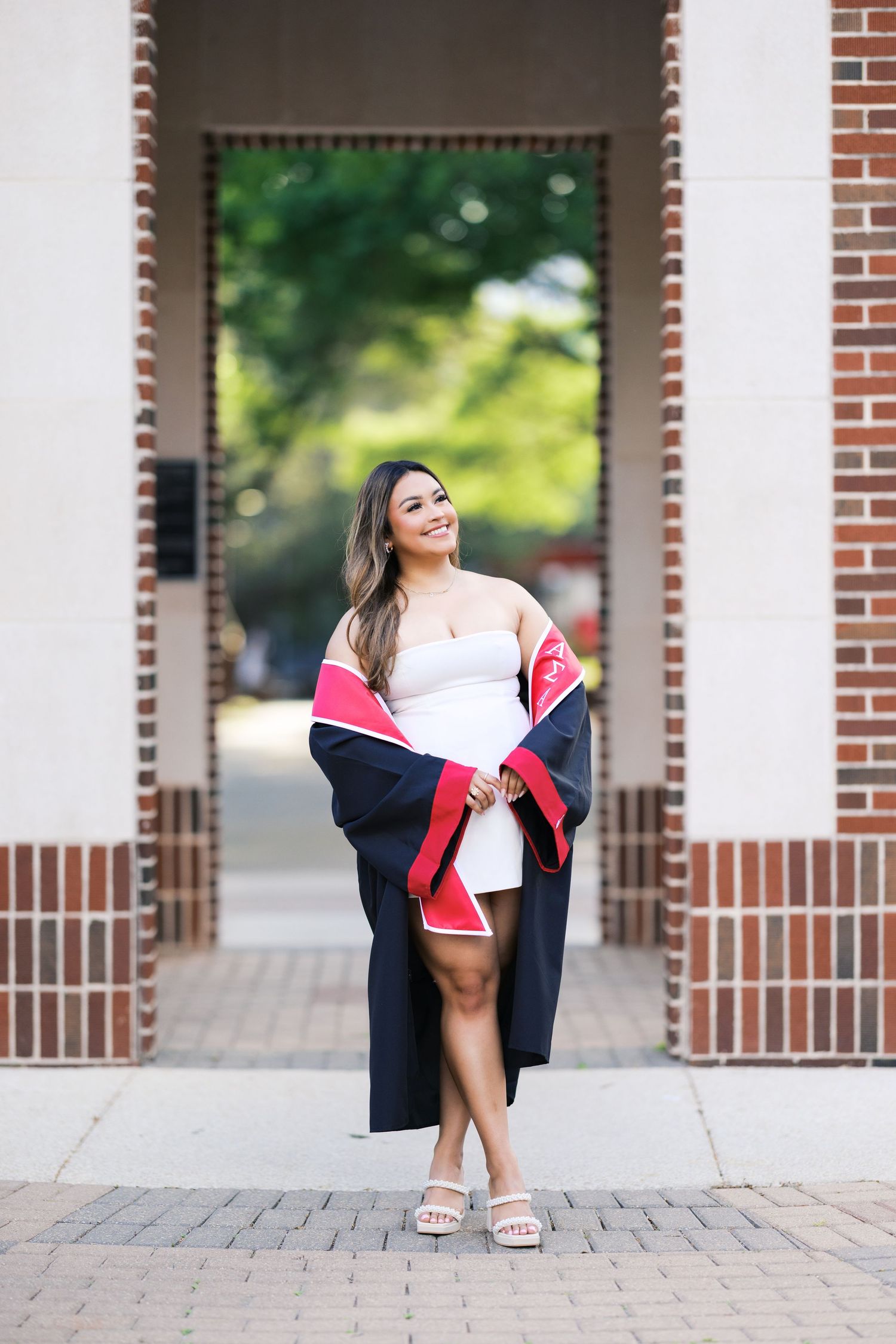 Smiling graduate poses in black and red graduation gown over white dress at brick archway.