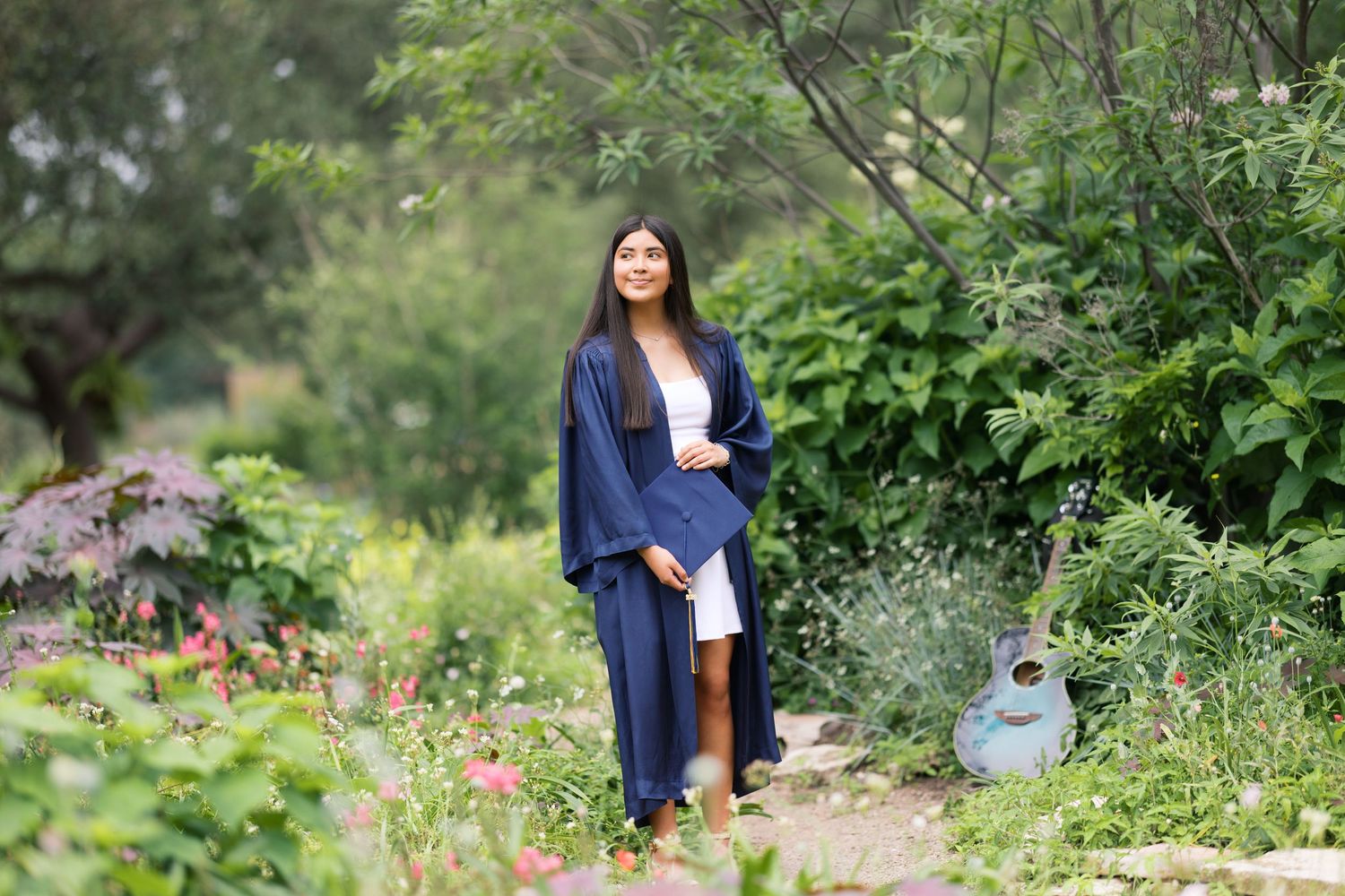 Young woman in navy graduation gown poses in lush garden with guitar, surrounded by green foliage and flowers.