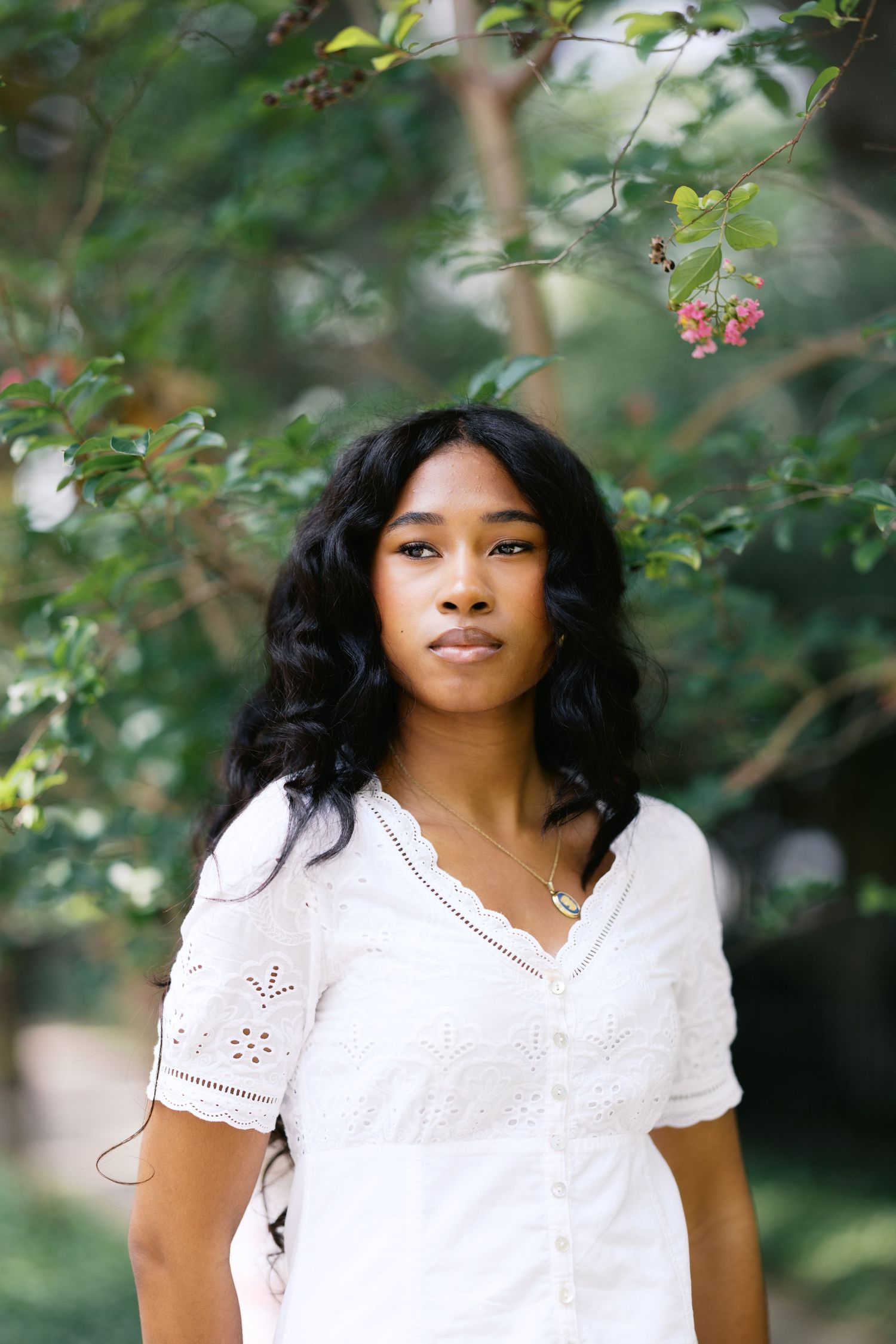 Young Black woman in white eyelet dress standing outdoors among green trees with pink flowers.