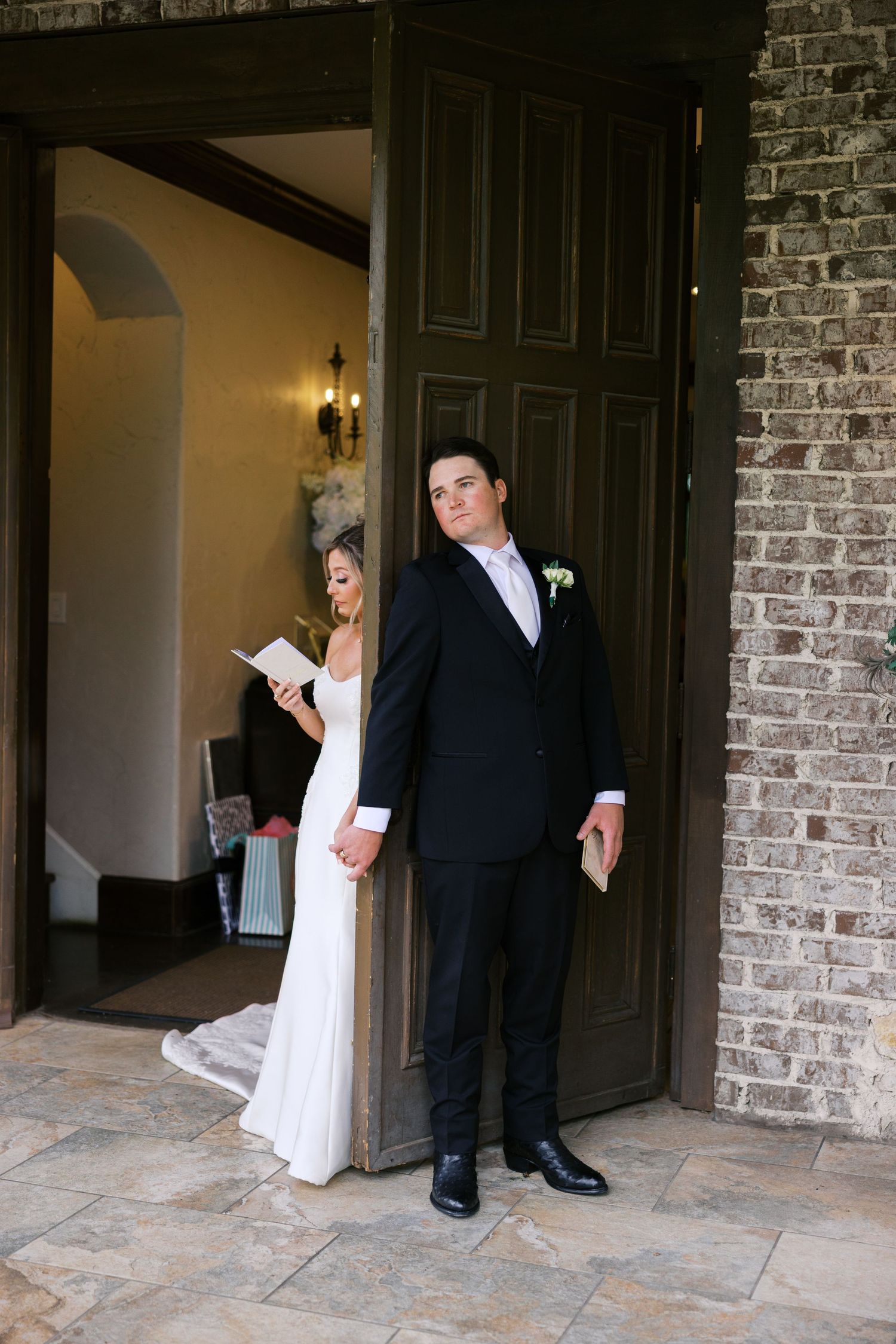 Bride reads vows while groom stands on opposite side of large wooden door during wedding first look.