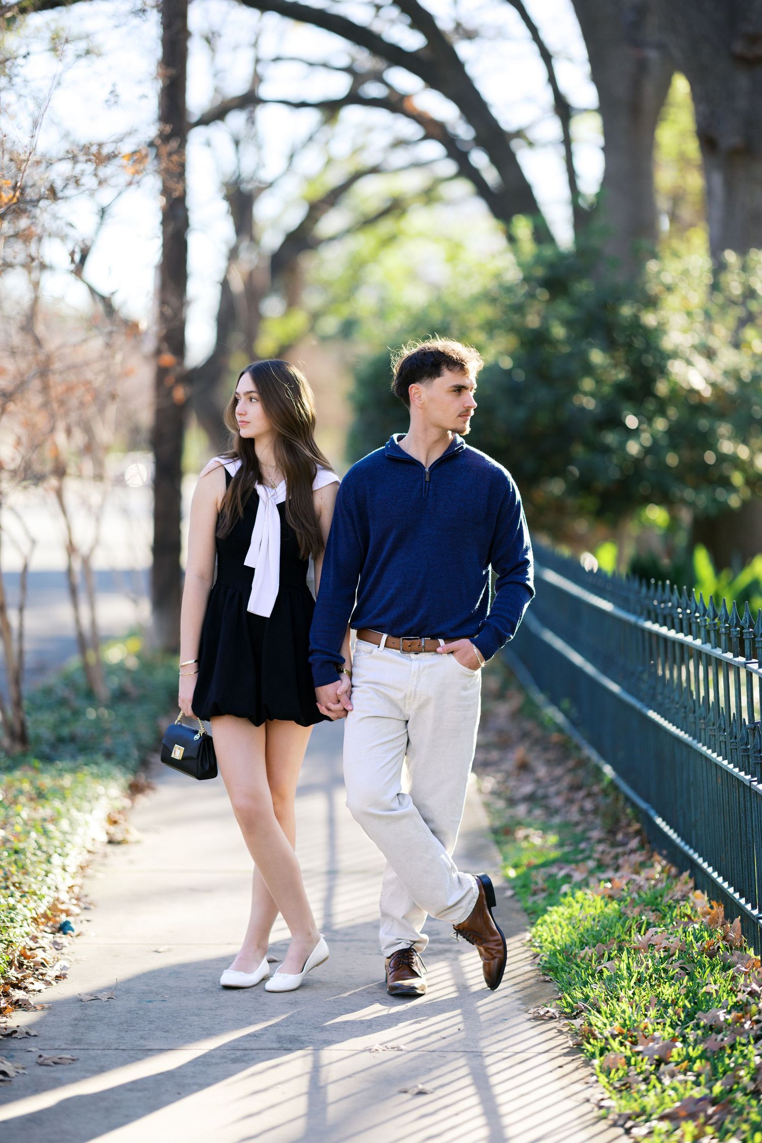 Stylish couple strolling along a park pathway beside a green fence, woman in black mini dress with white bow.