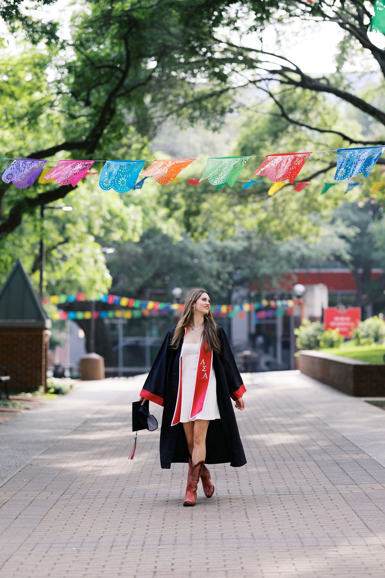 A graduate in black gown walks under colorful papel picado banners on a tree-lined campus path.