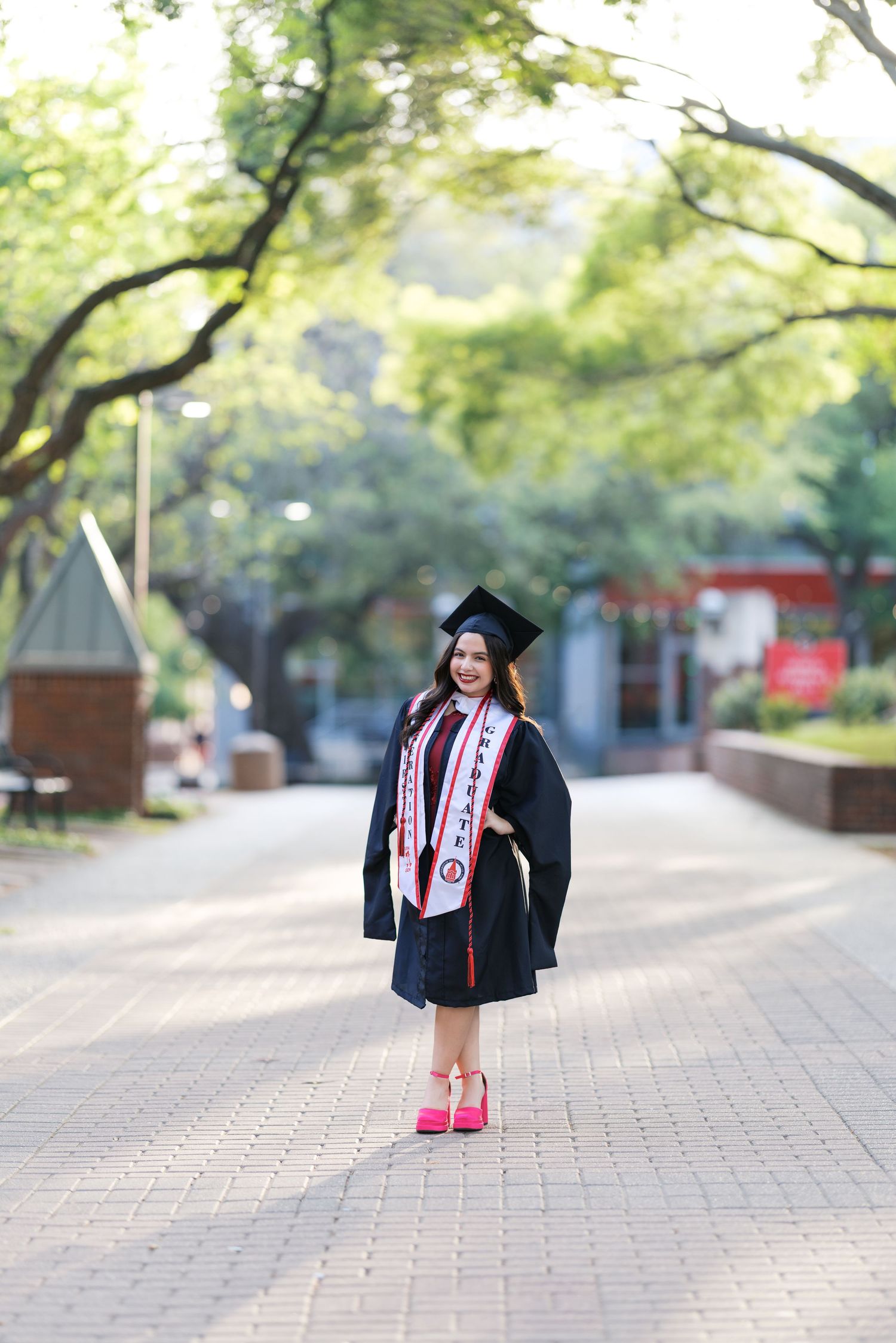 Graduate in black cap and gown with red stole stands on brick campus walkway lined with trees.