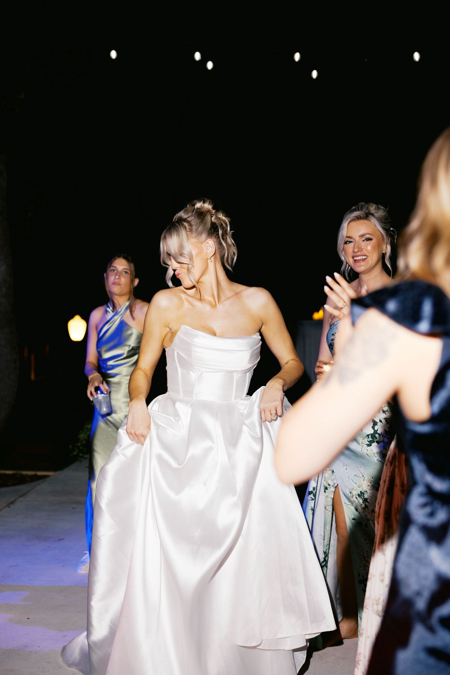 Bride in white strapless gown dancing joyfully at outdoor evening wedding reception with guests.