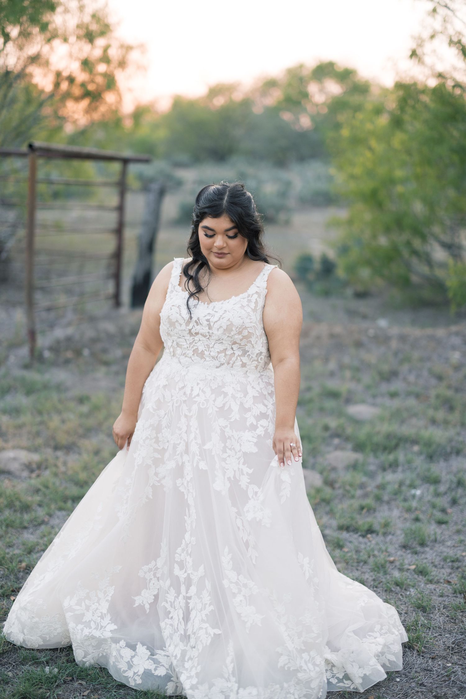 A beautiful bride in a lace wedding gown poses in an outdoor rustic setting at sunset.