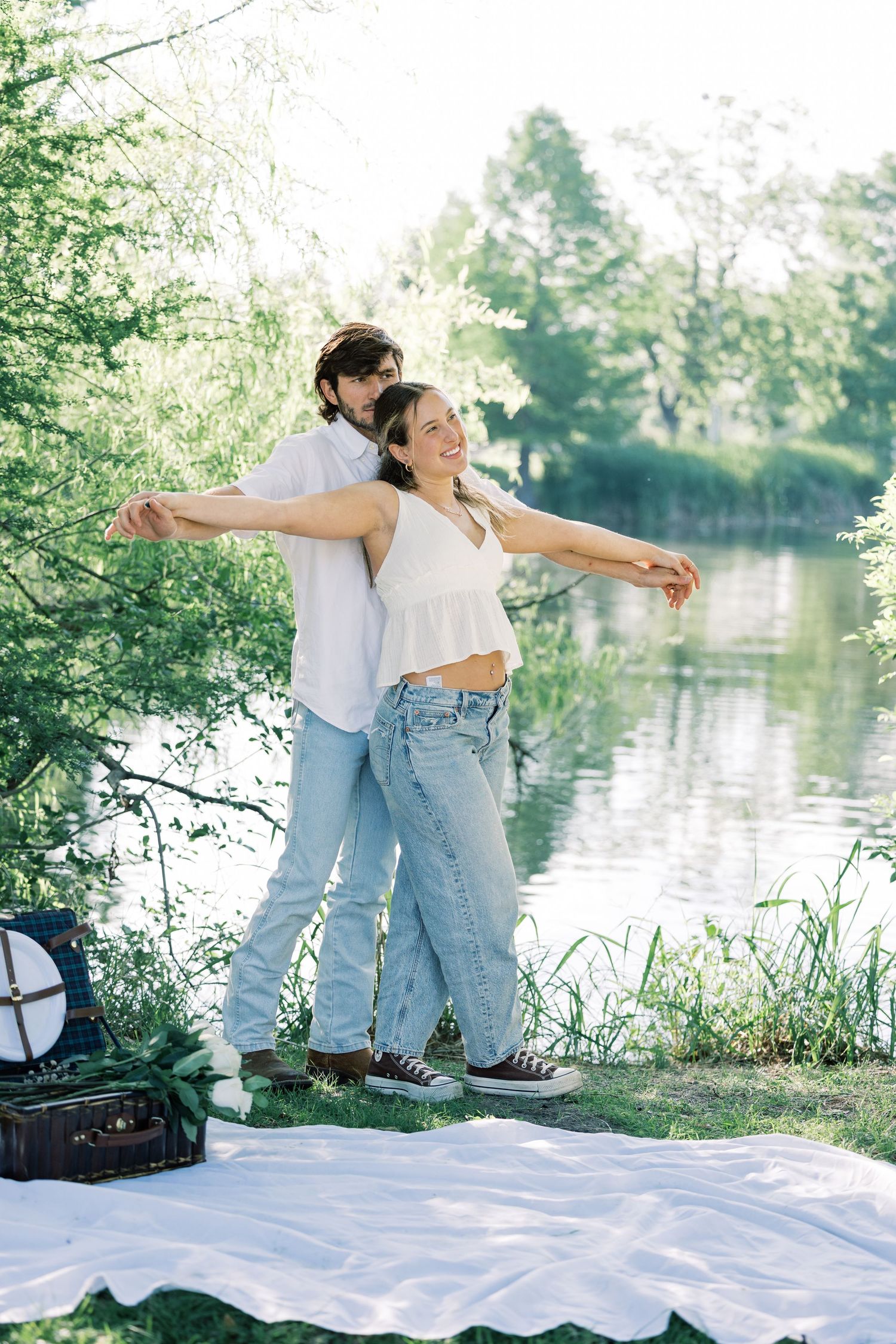 Two people strike playful poses with outstretched arms by a lakeside.