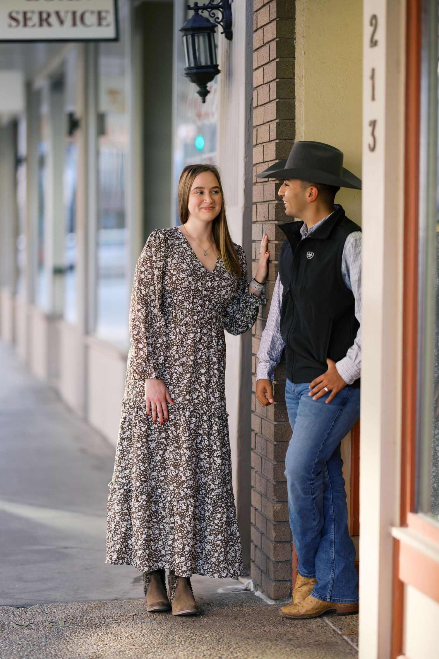 A couple posing against a brick wall with storefronts and a service sign visible.