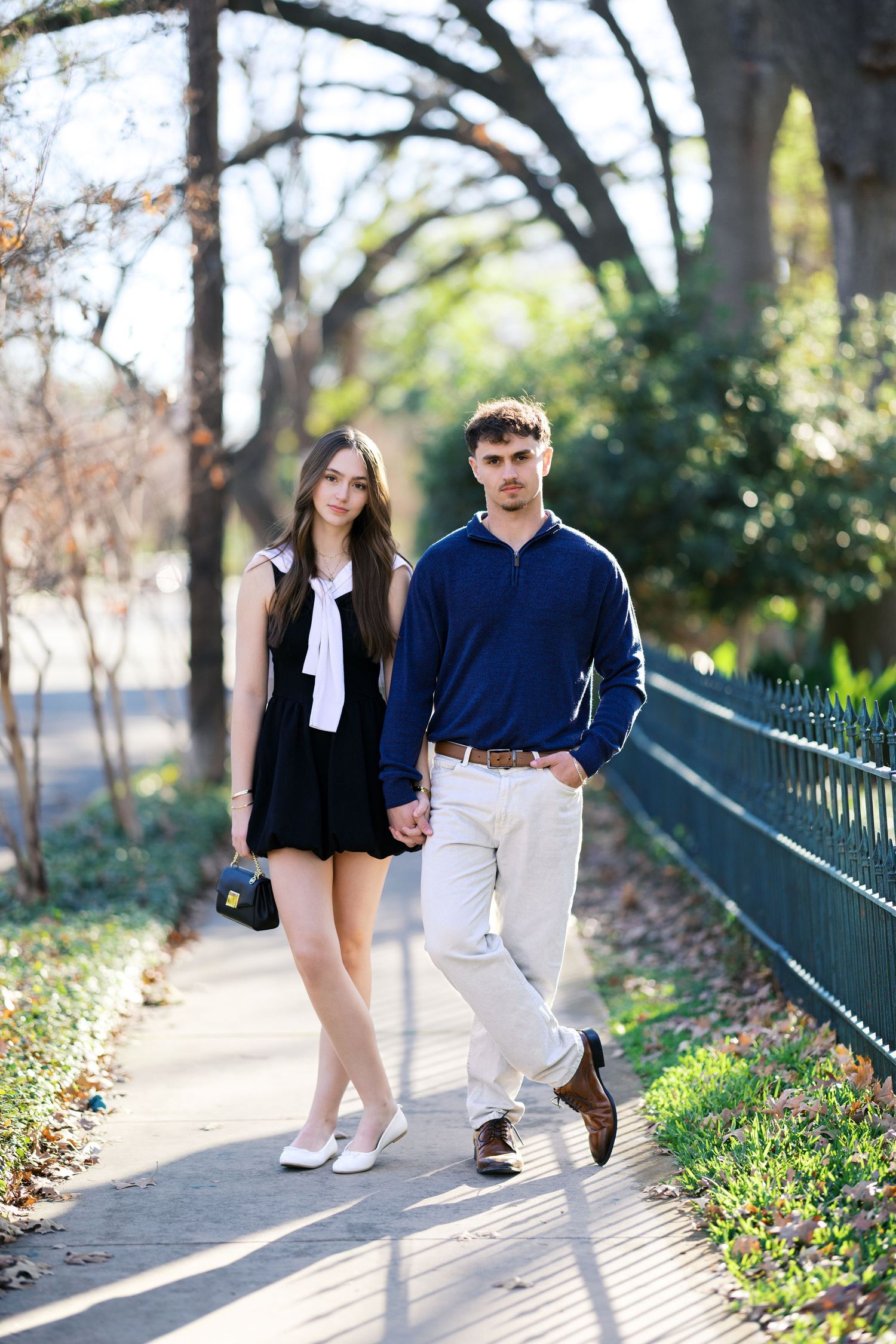 Young couple holding hands walking on a sunny park path, woman in black dress, man in navy sweater and white pants.