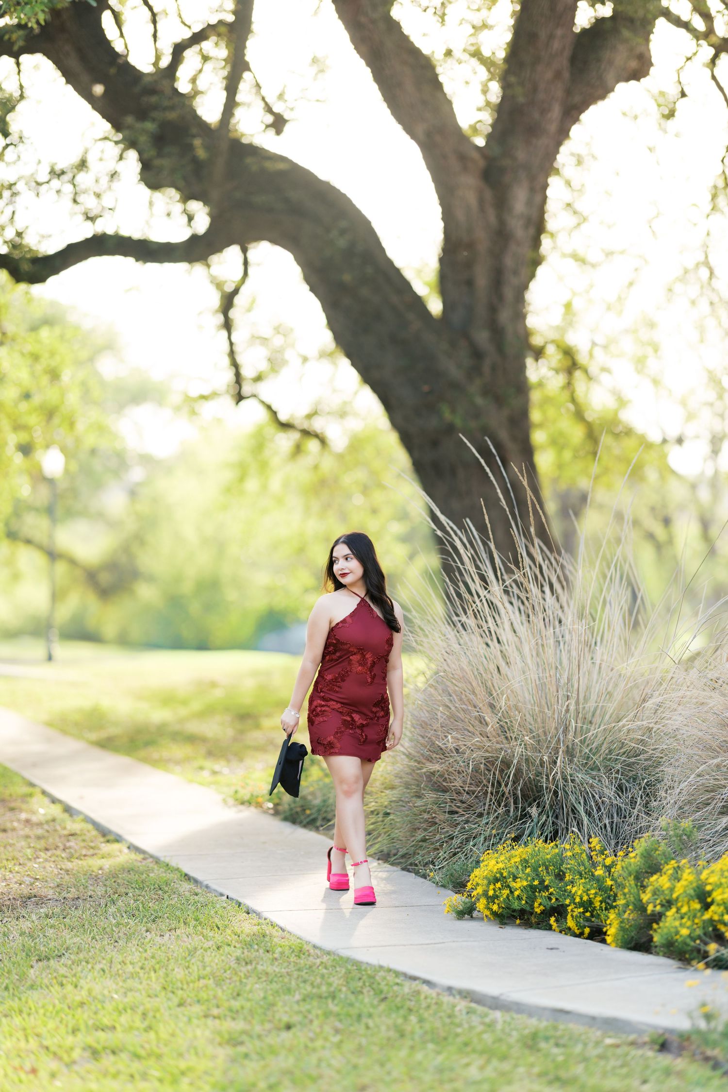 Young woman in burgundy mini dress and pink heels walks along garden path under large oak tree.