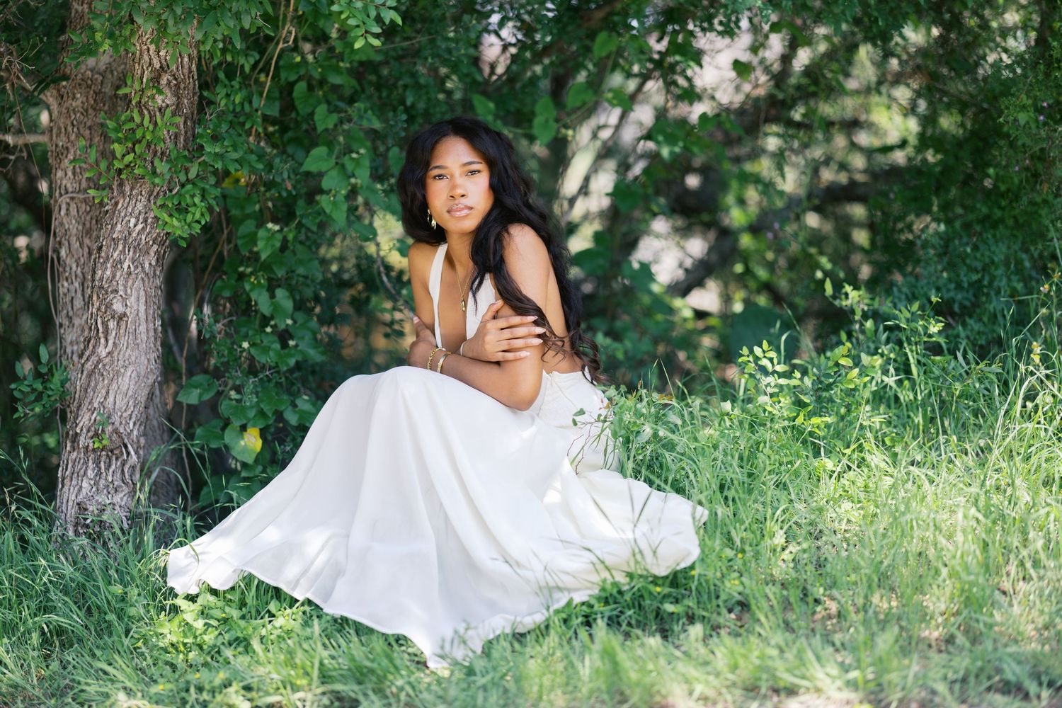 Young Black woman in flowing white dress sits gracefully in green meadow beside a large tree.