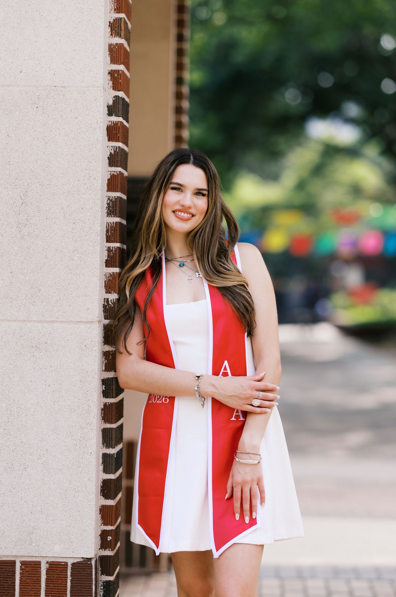 Smiling young woman in white dress and red graduation stole leaning against brick wall outdoors.