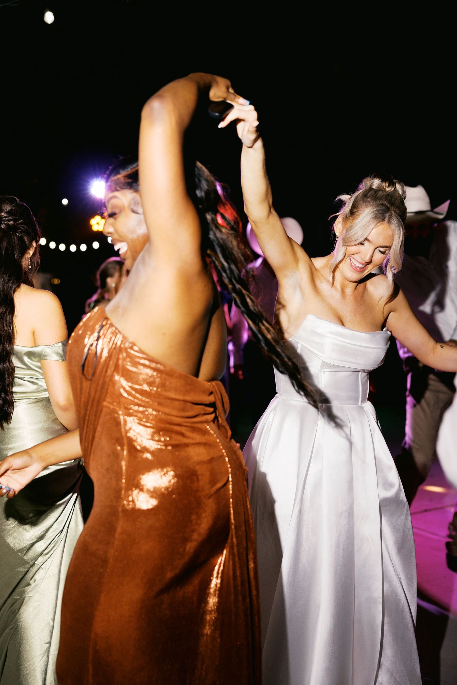 Bride in white strapless gown dancing joyfully with bridesmaid in copper dress at reception.