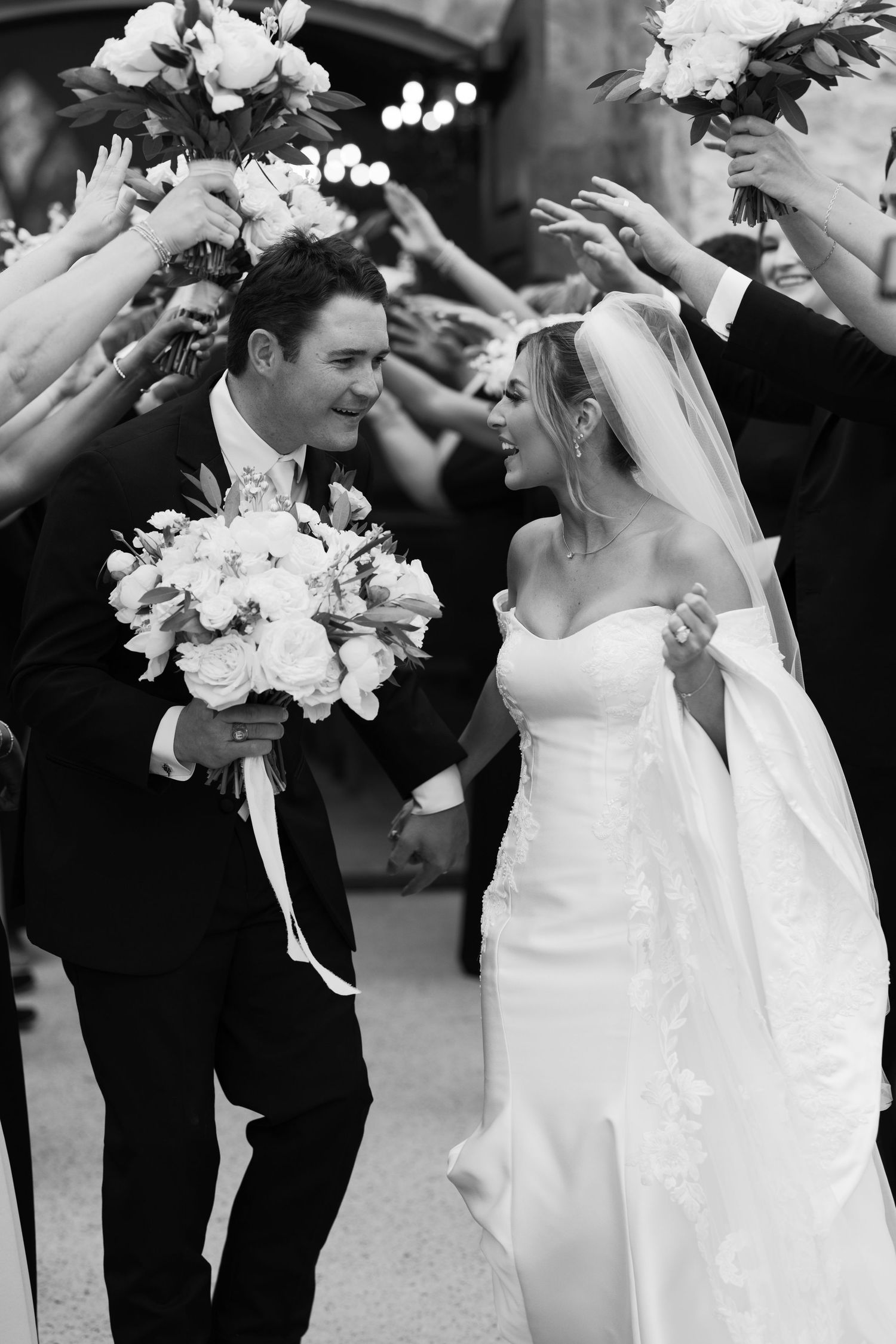 Black and white photo of joyful newlyweds walking through a celebratory arch of raised hands after the ceremony.