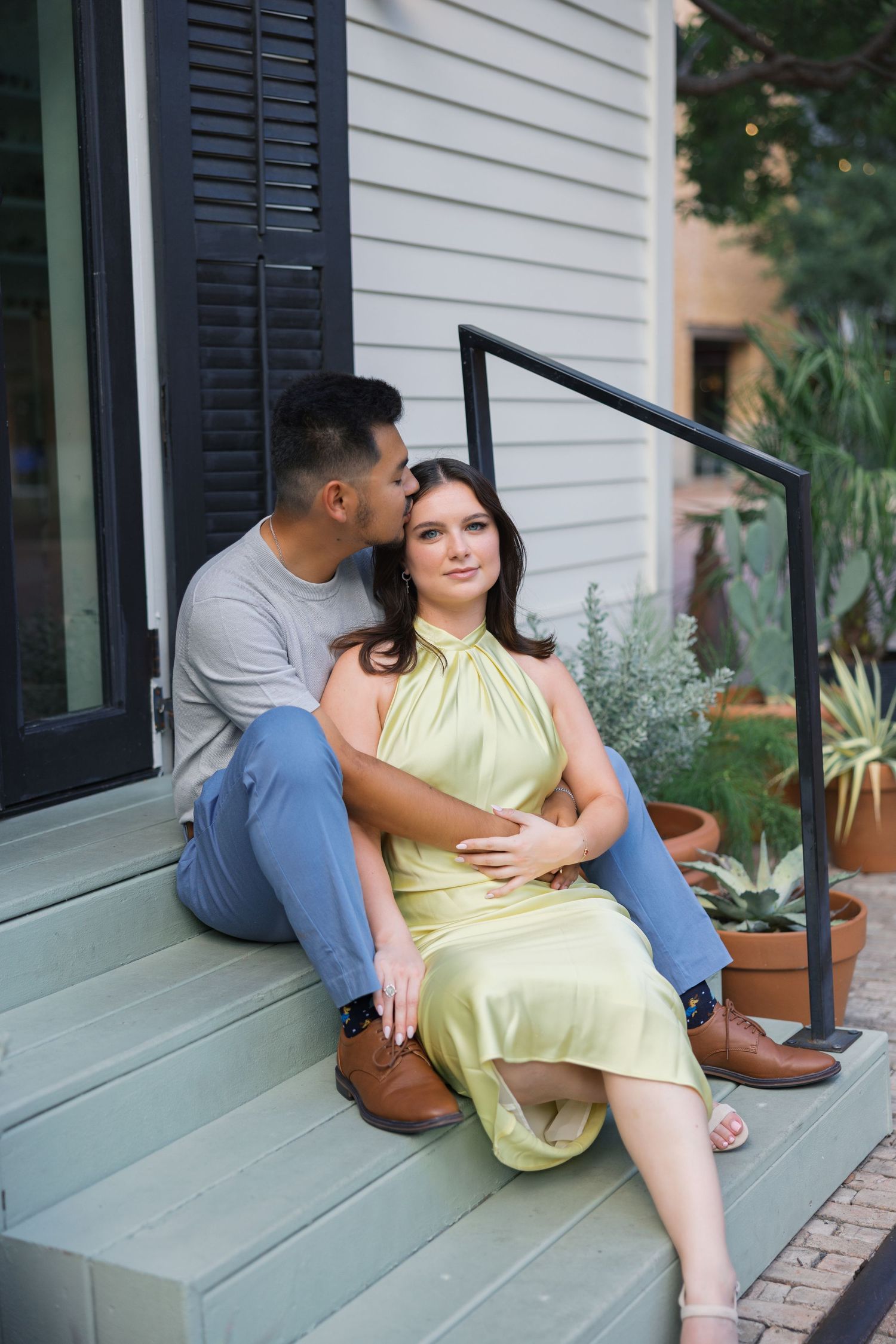 A couple sits together on outdoor steps with white siding and potted plants.