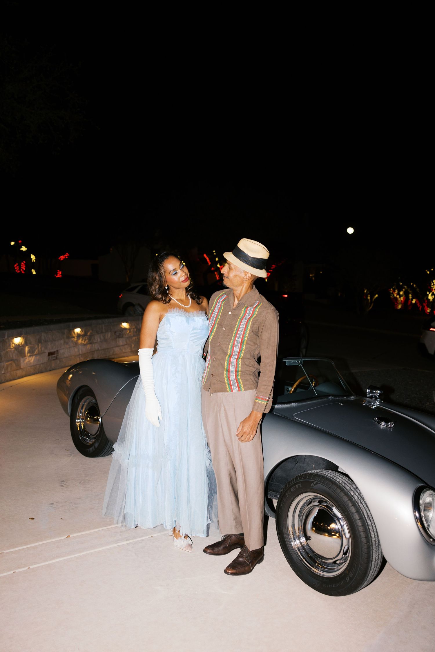 A couple in vintage formal wear poses beside a classic sports car at night.
