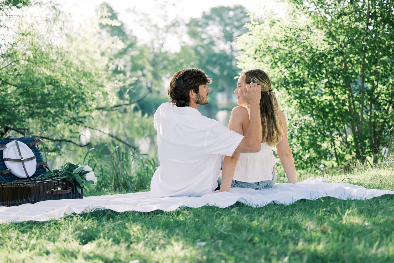 A couple shares an intimate moment on a white blanket in a sunlit park setting.
