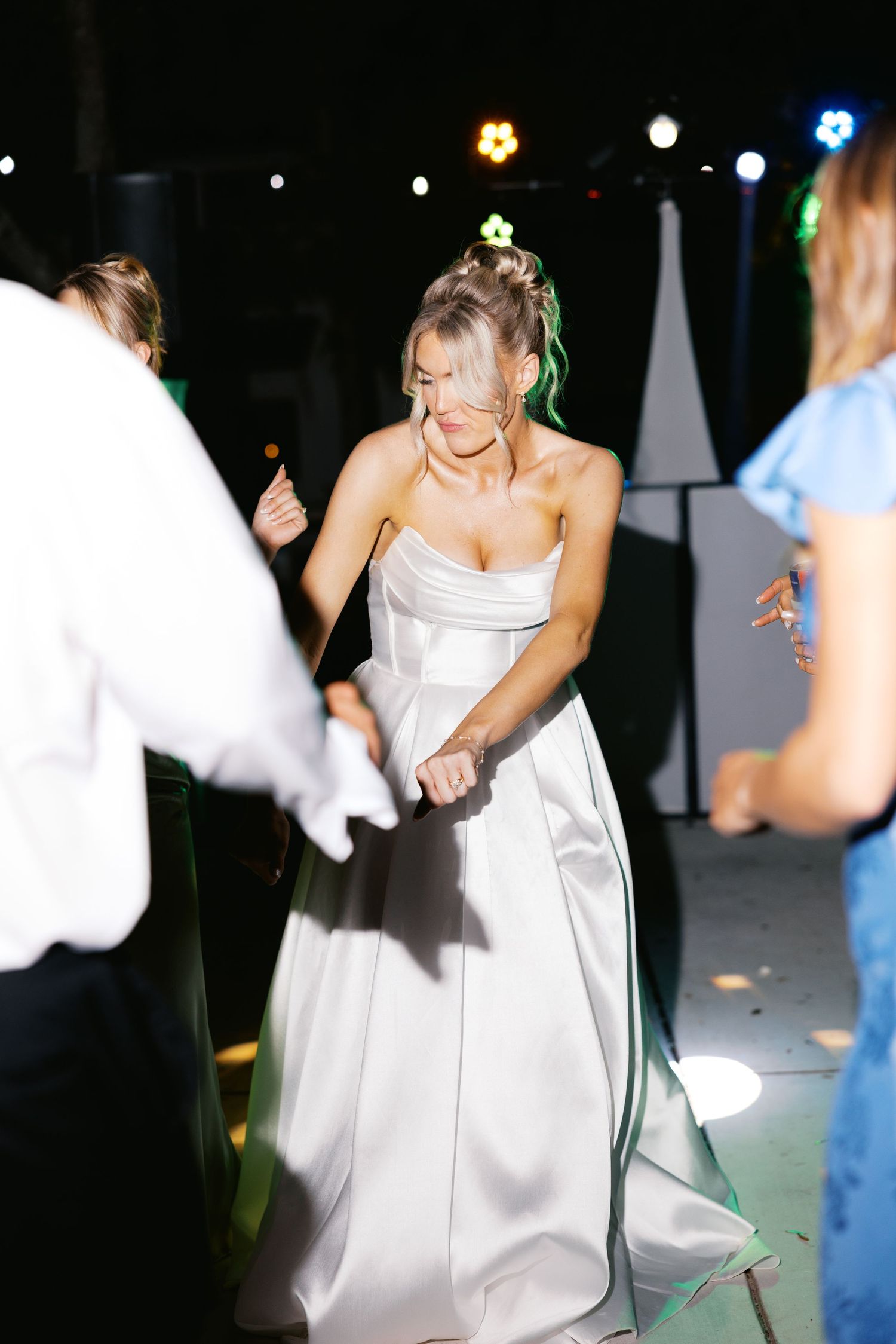 Bride in strapless white satin gown dances joyfully on outdoor dance floor at evening wedding reception.