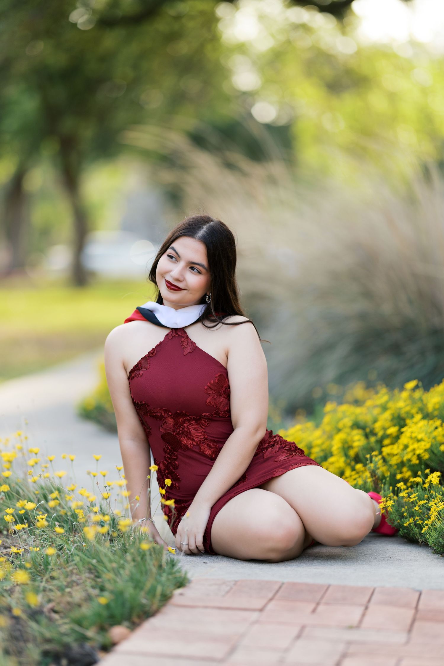 Young woman in burgundy lace dress kneeling on brick path surrounded by yellow flowers in a garden.